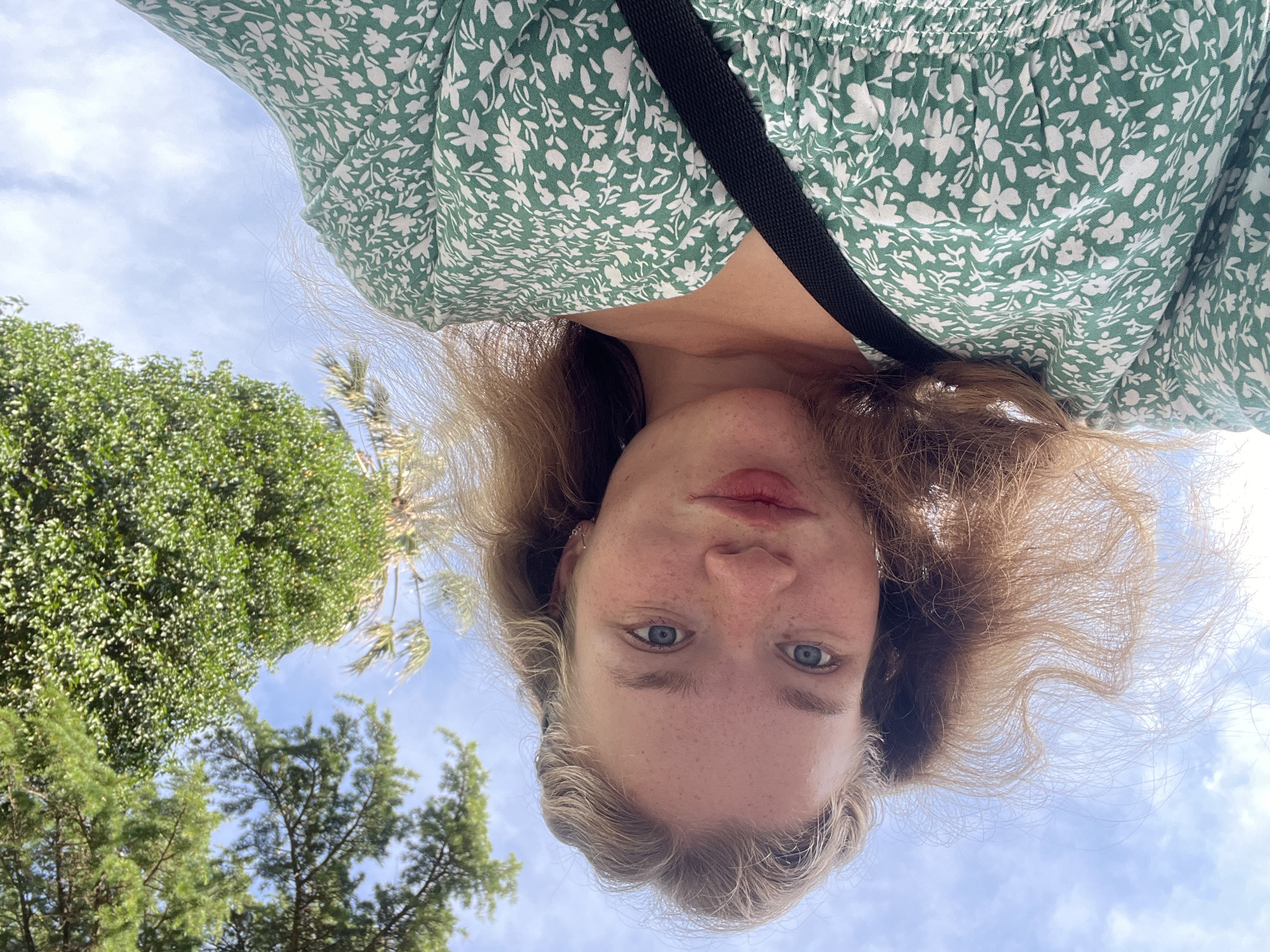 The image is an upside-down selfie of a fair-skinned person with freckles and blue eyes. They are wearing a green floral print top and a black strap is visible across their shoulder. The background includes trees and a cloudy sky. The exact location within Budva, Montenegro cannot be determined from the provided image.