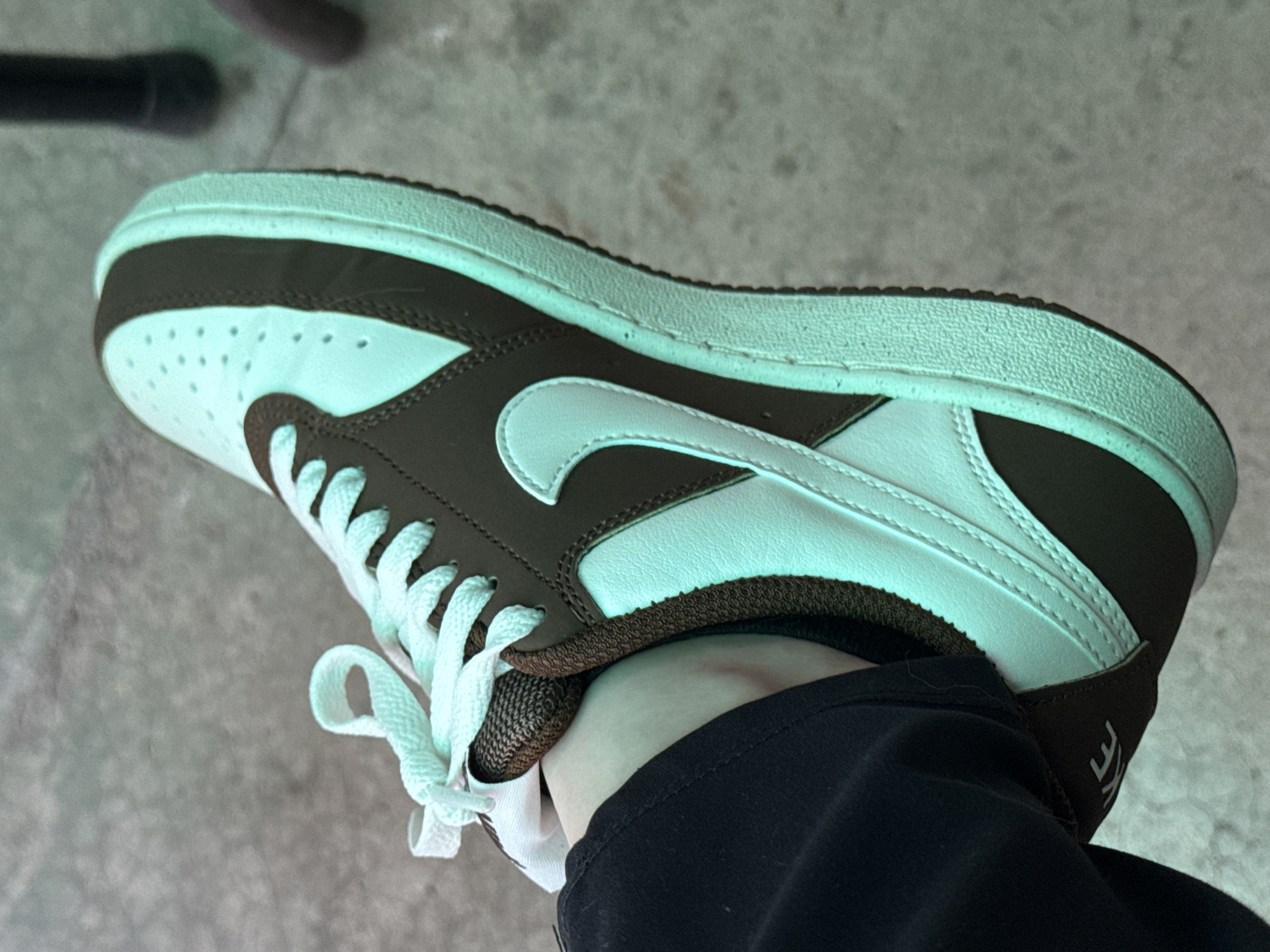 The image presents a close-up, high-angle view of a person's left foot wearing a low-top sneaker, positioned at a slight angle. The sneaker features a two-tone color scheme: dark brown and a light, muted blue-green (resembling mint or seafoam green). The light blue-green color covers the toe box (with visible perforations), central panels, the iconic Nike swoosh, tongue, laces, and the entire sole. Dark brown sections form overlays around the toe, eyelets, side panels, and the heel. The person's lower leg and ankle are partially visible, covered by a black garment, likely pants or a sock. A fragment of text, "KE", is visible in white on a black section of the shoe's heel, indicating the brand "NIKE". The background is a textured, plain concrete or paved surface. The lighting appears natural and diffuse, suggesting it is daytime. The image captures a casual moment of the sneaker being worn. This scene takes place in Govedare, Bulgaria.
