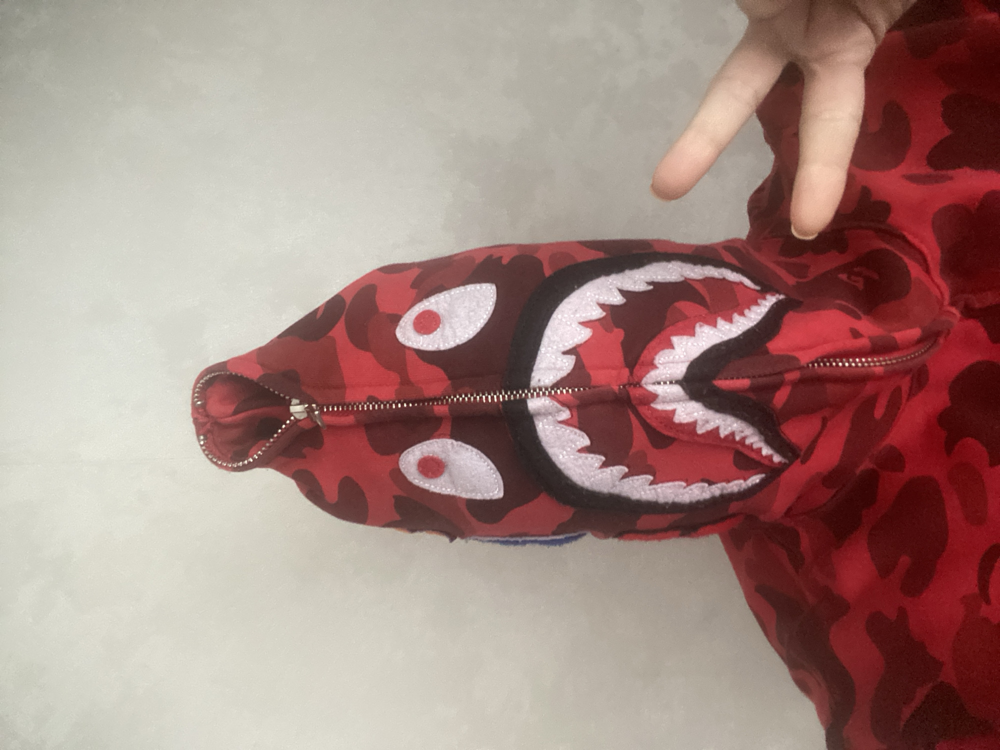 A red camouflage hooded sweatshirt with a shark-like design on the hood is lying on a light gray surface. A hand is visible in the upper right corner, making a peace sign. The zipper of the hoodie is partially open. The location is not discernible from the image and therefore the location in Sochi, Russia cannot be determined.