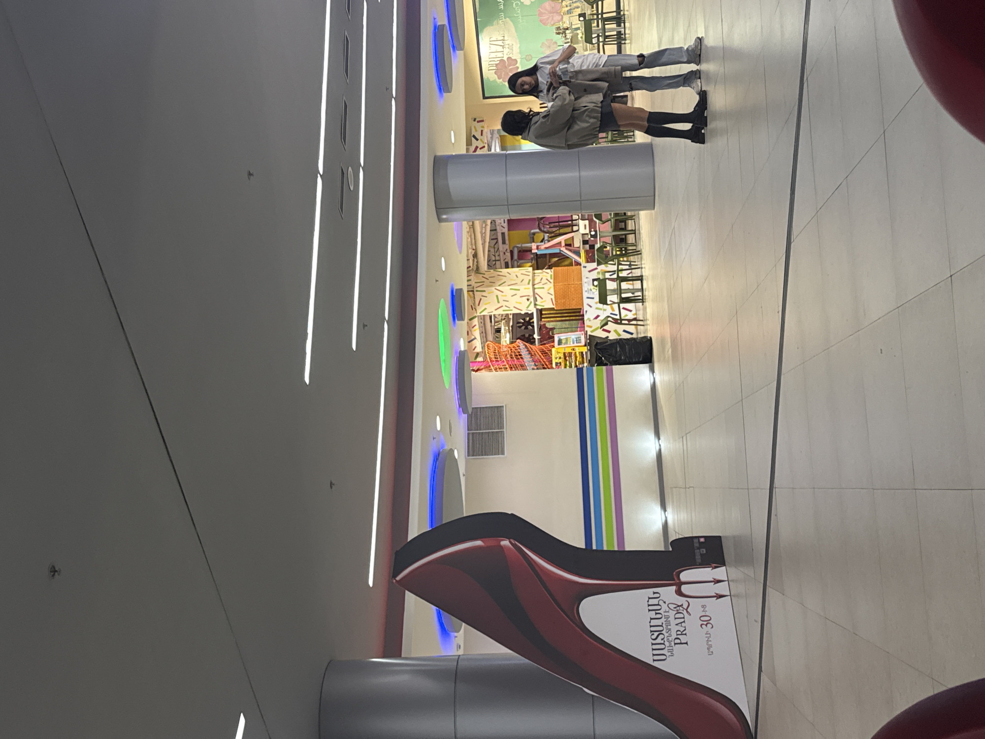 The photograph shows an indoor scene in Yerevan, Armenia. A large advertisement for Prada, featuring a high-heeled shoe with devil-like horns, is visible in the foreground. In the background, two people stand near what appears to be a children's play area. One person is wearing ripped jeans and a white jacket, while the other is wearing a grey jacket with knee-high black socks. The environment suggests a shopping mall or similar commercial space, with colorful decor and architectural elements.