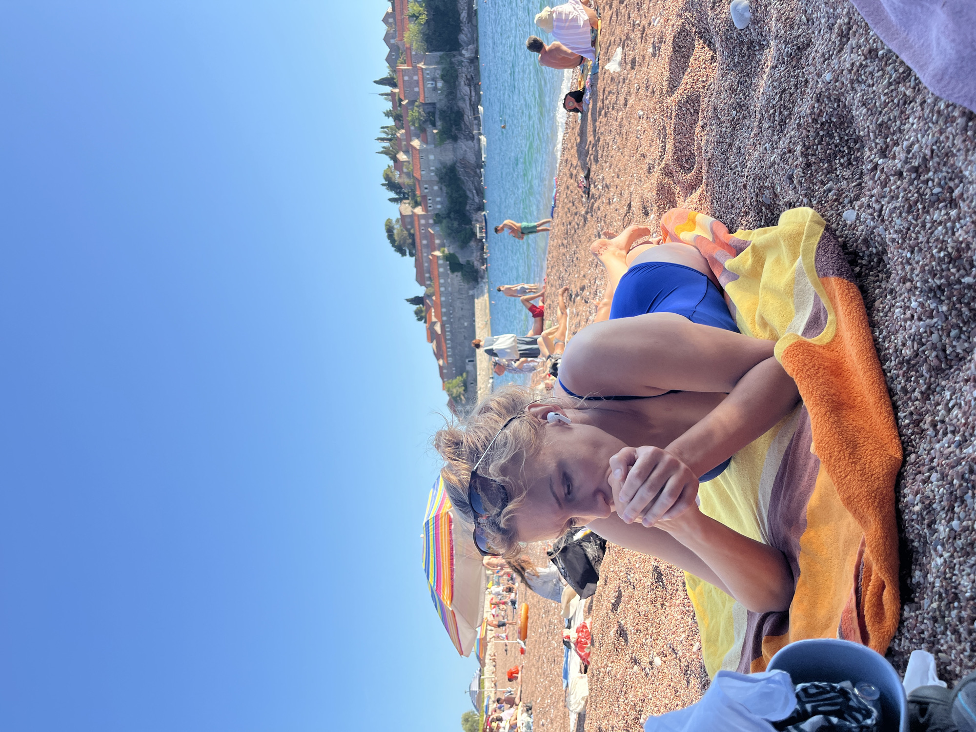 The image captures a sunny beach scene in Rađenovići, Montenegro, with the fortified island of Sveti Stefan prominently visible in the background. In the immediate foreground, a blonde woman with curly hair, wearing a blue swimsuit and white wireless earbuds, lies prone on a striped beach towel (orange, yellow, and brown/purple). Her hands are clasped together near her mouth, and sunglasses rest on top of her head. To her left, partially visible, are a white garment and a blue container. The beach consists of reddish-brown and grey pebbles.
In the midground, numerous other individuals are present on the beach and in the clear blue-green water. Several beach umbrellas provide shade, including a large, rainbow-striped one to the left of the main subject. People are engaged in typical beach activities such as sunbathing, standing, and swimming. Small boats are visible in the water near the island. The background is dominated by the distinctive architecture of Sveti Stefan, an island covered with historic stone buildings featuring red-tiled roofs and interspersed greenery, situated across the calm sea under a clear blue sky.