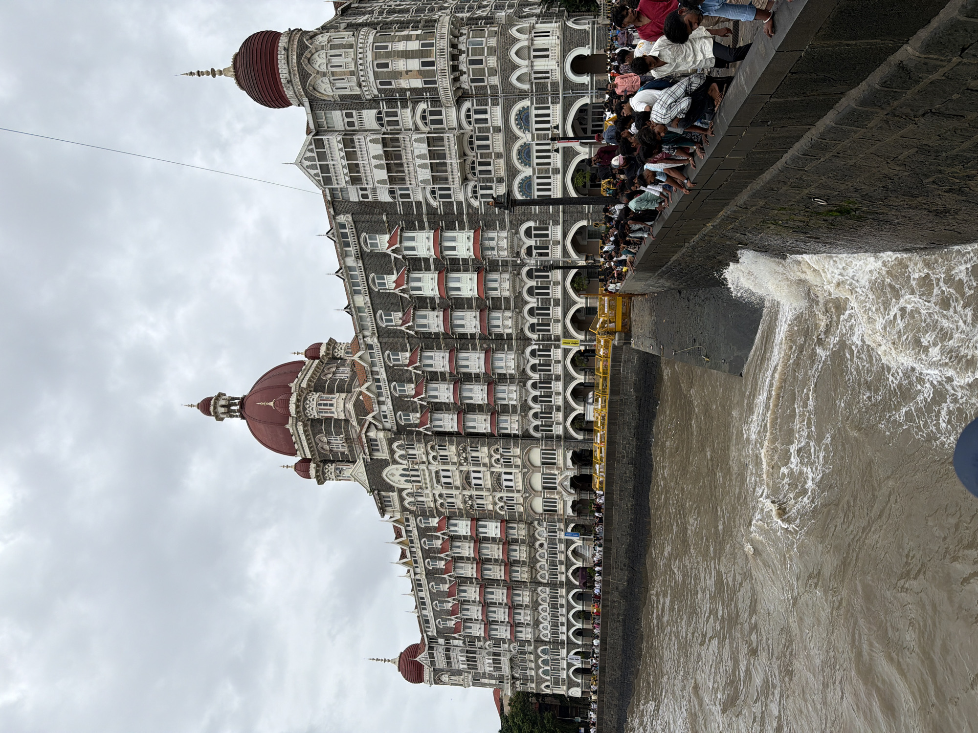 A wide shot captures the iconic Taj Mahal Palace Hotel in Mumbai, India, on a cloudy day. The grand hotel, with its distinctive red domes and intricate architecture, stands tall against a gray sky. A crowd of people are gathered along a stone embankment, leaning over to observe the choppy, brown water that laps against the wall. Waves crash against the embankment, creating white foam. The hotel's facade is adorned with numerous windows and decorative elements, including arched doorways on the ground level. People are dressed in casual clothing, and some are looking at their phones. The scene suggests a popular tourist spot, possibly a waterfront promenade, with the hotel serving as a prominent landmark.