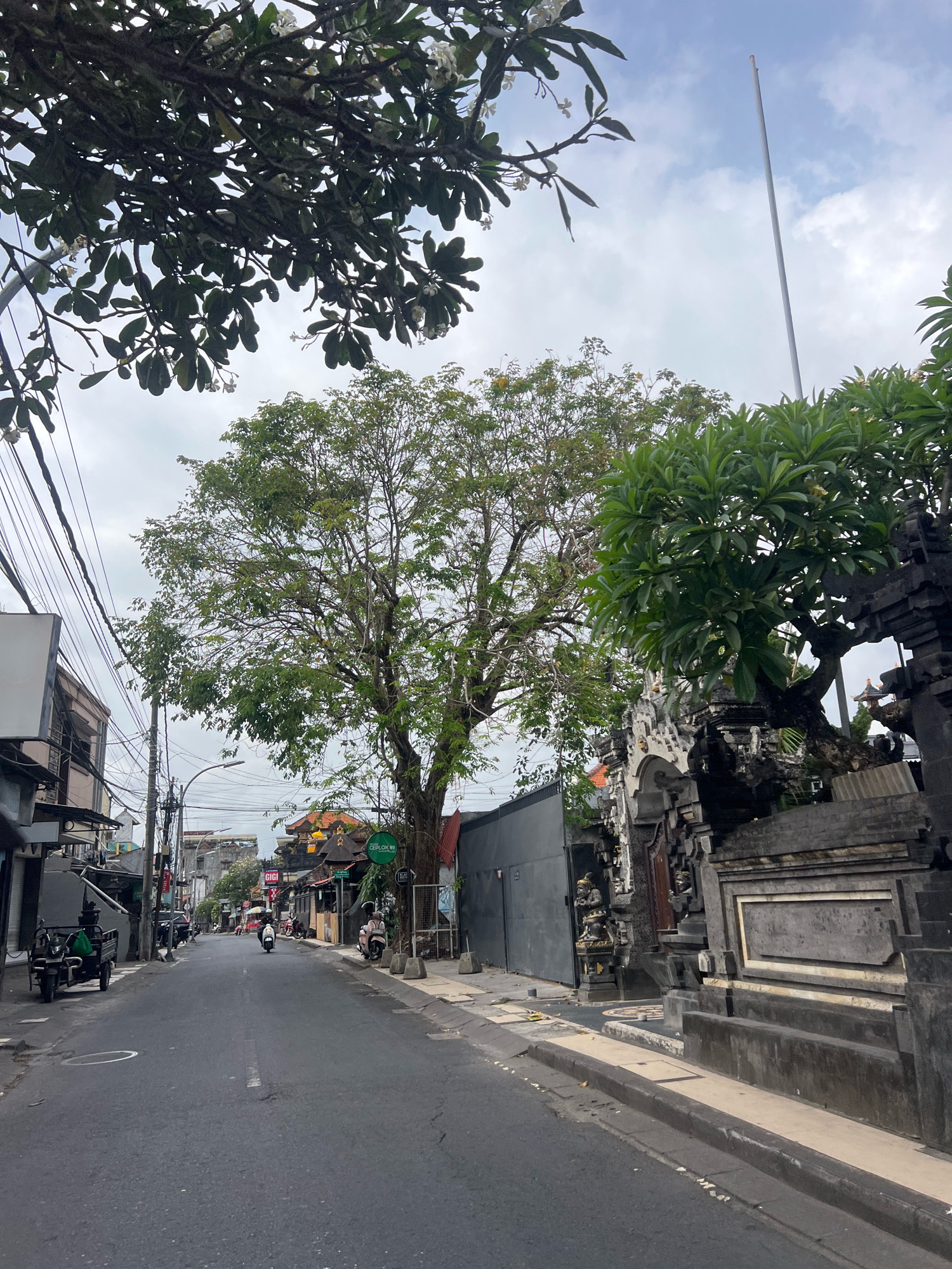 The image depicts a street scene in Kuta, Indonesia. A paved road runs through the center, lined with buildings on both sides. A large tree with green foliage dominates the right side of the frame, casting shadows on the road. In the foreground, on the right, there's an intricately carved stone gate, possibly part of a temple or traditional Balinese architecture. Several motorcycles are parked along the side of the road, and one motorcycle is visible on the street, ridden by a person. Power lines crisscross the sky above the buildings. The sky is overcast, with scattered clouds. The overall impression is of a typical urban street in a tropical climate.