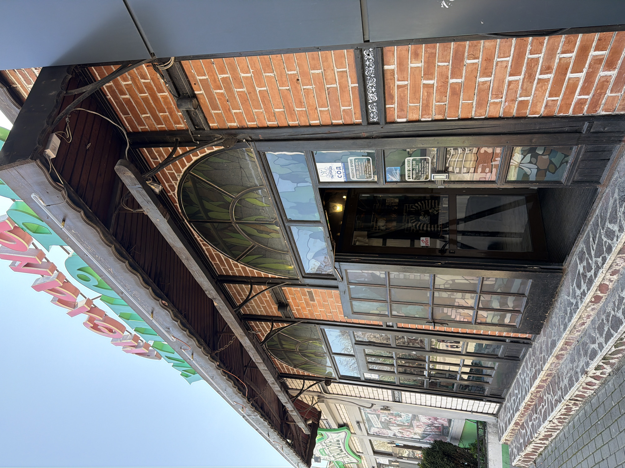 The image displays an angled exterior view of a commercial building at 24 General Popov Street in Varna, Bulgaria. The facade features reddish-brown brickwork with white mortar, interspersed with dark-painted wooden beams and trim. A dark wooden canopy, supported by black metal brackets, extends over the entrance, revealing exposed electrical wiring and a fluorescent light fixture underneath. Above the canopy, a large illuminated sign with raised pink and green letters partially spells "Механа" (Mehana).
Three steps, surfaced with irregular dark and reddish-brown stone pieces, lead up to the entrance. The entrance consists of double doors, framed in dark wood, with multiple small glass panes. Stained-glass elements in greens, blues, and browns are present in the semicircular arched tops and lower door sections. The right door is slightly ajar, revealing a dark interior and an internal light source.
Several white signs with black text are affixed to the right door panel: one reads "СОД" (SOD) and another states "РАБОТНО ВРЕМЕ" (Working Hours) as "09:00 - 23:00 every day," followed by the address "Varna, 24 Gen. Popov Str." and a phone number. A small red sign with white text is partially visible on the right door frame. To the immediate right, the brickwork transitions to a dark gray vertical panel. To the left, an adjacent facade with lighter brickwork and a partially visible green and white sign is present. The foreground is paved with grey rectangular pavers. The sky is clear and blue. No individuals are distinctly visible.