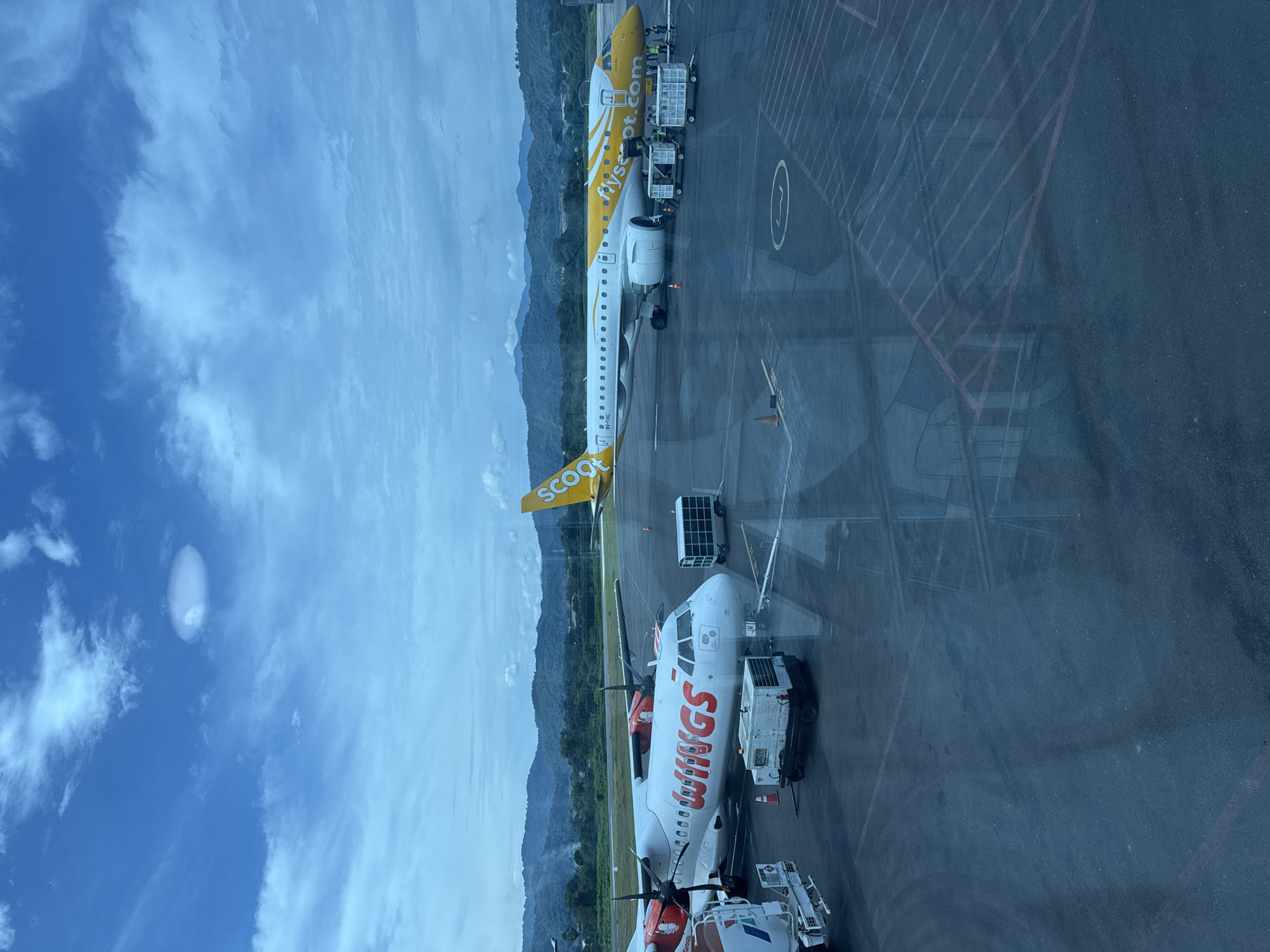 The image shows an airport scene. Two airplanes are visible: one branded "SCOOT" in yellow and white, and another "WINGS" in red and white. They are parked on the tarmac, likely being serviced or prepared for flight. There are vehicles around both planes that appear to be baggage carriers or service vehicles. The tarmac is marked with lines and a circular area. In the background, mountains are visible beneath a cloudy sky. The location is in the city of Unknown, Unknown. The image was likely taken from inside the terminal building, evidenced by the glass reflection in the lower right area.