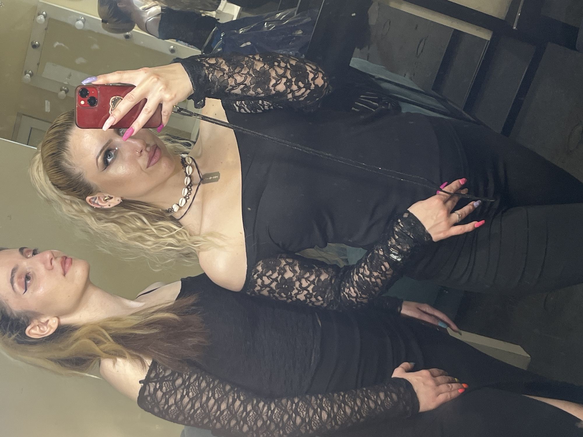 A selfie taken in a mirrored vanity area, possibly a dressing room. Two fair-skinned women are visible. One, with blonde hair, holds a red smartphone taking the picture. She wears a black off-the-shoulder top with lace sleeves and a necklace featuring shells and a razor blade pendant. Her nails are painted in various colors. The other woman, with brown and blonde hair, is positioned beneath the first. She also wears a black top with lace sleeves. The background reveals vanity lighting, a mirror, and assorted items suggesting a backstage or dressing room environment. The location is not explicitly identifiable as Plovdiv, Bulgaria from the image alone.