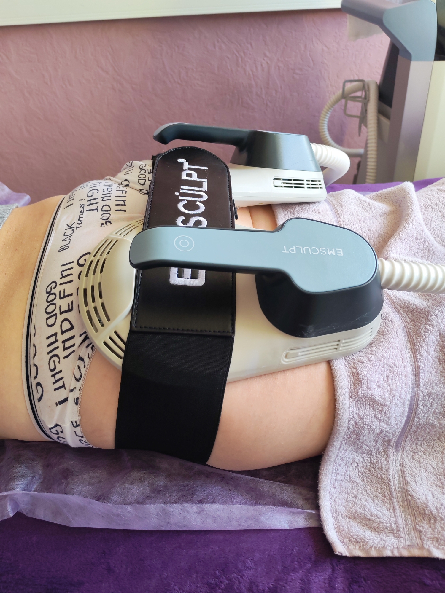 The image shows a person lying on a purple-covered table with an Emsculpt machine attached to their abdomen. The machine has a black strap securing it. The person is wearing underwear with the words "GOOD NIGHT! INDEFINI BLACK INS" printed on them. A light purple towel is draped beside the person. In the background, a medical device with a hose is visible. The image likely depicts a cosmetic procedure being performed. The location is Chaikovsky, Russia.