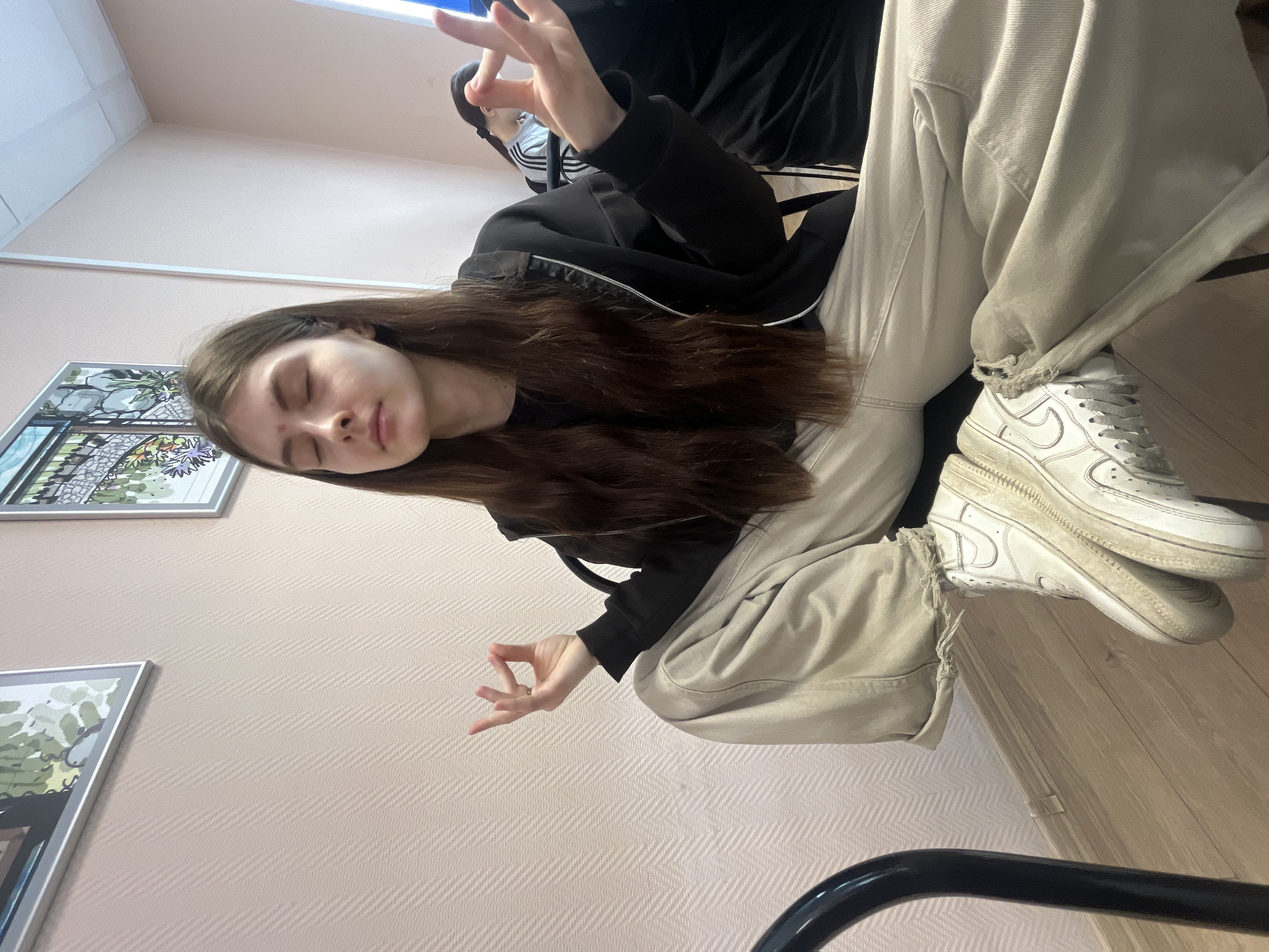 A young woman with long, dark brown hair is depicted sitting and appearing to meditate or be in a state of deep relaxation. She is wearing a black zip-up hoodie or jacket, light beige loose-fitting trousers that are slightly distressed at the ankles, and white Nike sneakers that show some wear. Her eyes are closed, and her face is serene, with a small blemish visible on her forehead. Her hands are positioned in a mudra gesture, with the thumb and index finger of each hand touching.
The setting is an indoor room. The walls are covered in light pinkish-beige wallpaper featuring a subtle diagonal striped pattern. Two framed pictures with abstract or stylized landscape illustrations in silver frames hang on the wall. The floor is composed of light-colored wooden planks. Parts of a black chair are visible in the foreground and background, suggesting she is seated on one. Another person is partially visible in the background, also seated.
The lighting suggests an indoor, artificial environment. The subject's pose, closed eyes, and hand gestures convey a sense of peace, concentration, or tranquility. No visible text is present in the image.