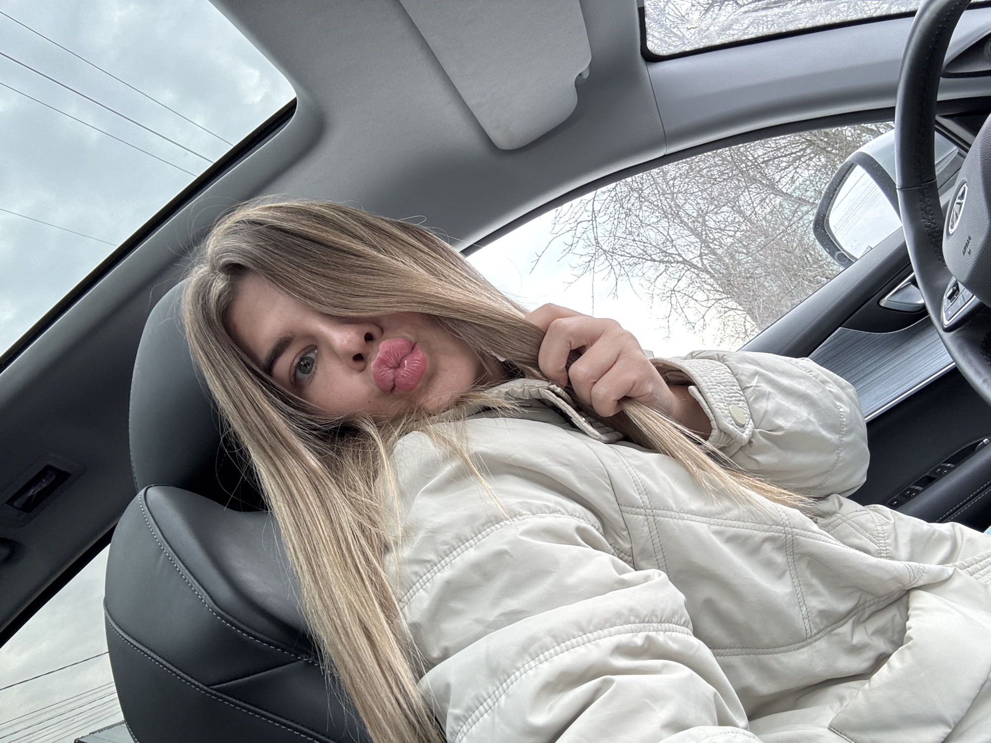 A young, fair-skinned woman is seated in the driver's seat of a car, likely taking a selfie. She has long blonde hair, pursed lips, and is wearing a light-colored, quilted jacket. She is holding a strand of her hair with her right hand. The car has a sunroof and a black leather interior. The outside environment appears overcast with bare trees visible through the car window. The image was taken in Chaikovsky, Russia.