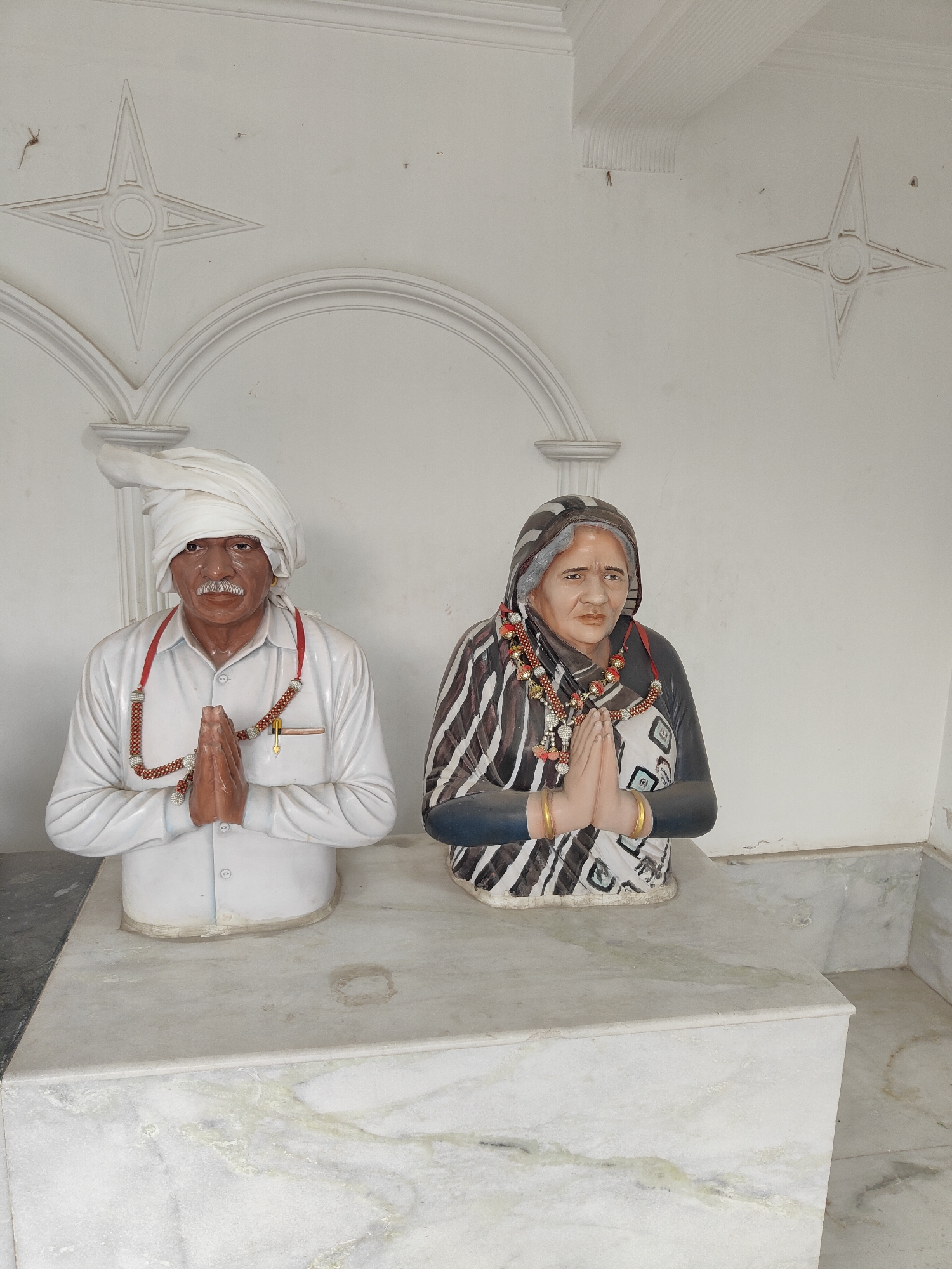 This is a medium shot of two plaster statues, a man and a woman, positioned side-by-side on a white marble platform. The man is on the left, wearing a white turban, a white shirt, and a red beaded necklace with a yellow pen tucked into his pocket. He has a mustache and his hands are clasped in front of him in a gesture of greeting or prayer. The woman is on the right, wearing a dark and striped headscarf, a patterned garment, and similar red beaded necklaces, with gold bangles on her wrists. Her hands are also clasped in front of her. Both statues appear to be lifelike in size and are placed in front of a white wall with subtle architectural details, including arched sections and star-shaped motifs. The scene is lit by overhead light, casting soft shadows. The marbles of the platform show some staining and wear. The image was taken outdoors, likely in a public or religious space in Gurgaon, India, based on its cultural depiction of traditional attire and greetings.