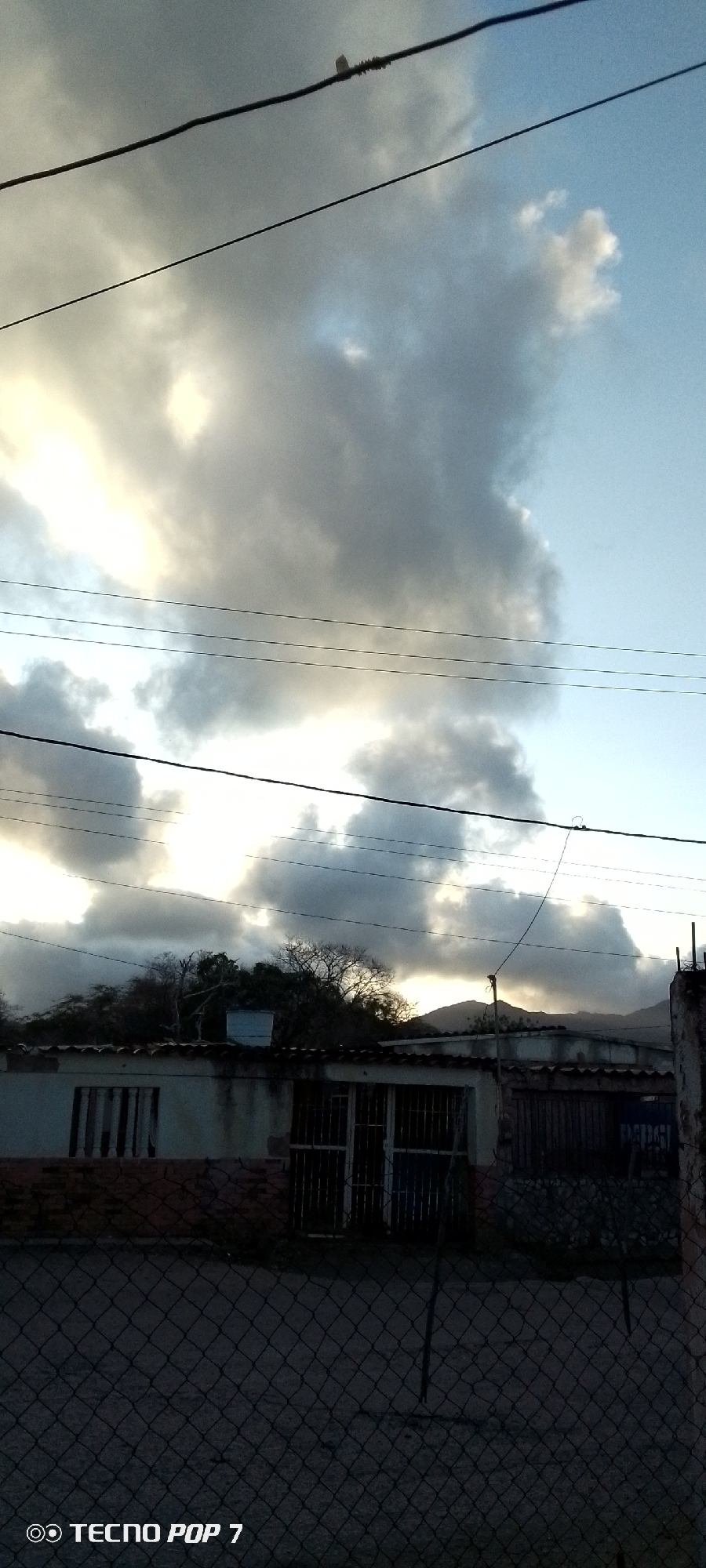 The image, taken in Altagracia, Venezuela, shows a cloudy sky with telephone wires running across the upper portion of the frame. Below, a low, single-story building with multiple sections is visible. The building has a mix of window types, including some with metal bars. Vegetation is growing behind the building. A chain-link fence runs across the foreground, partially obscuring the view. In the background, a mountain range is visible. The "TECNO POP 7" watermark is present in the lower-left corner. The overall lighting suggests either sunset or sunrise. There are no people visible. The image appears to be taken from behind the fence, looking towards the building and the sky.