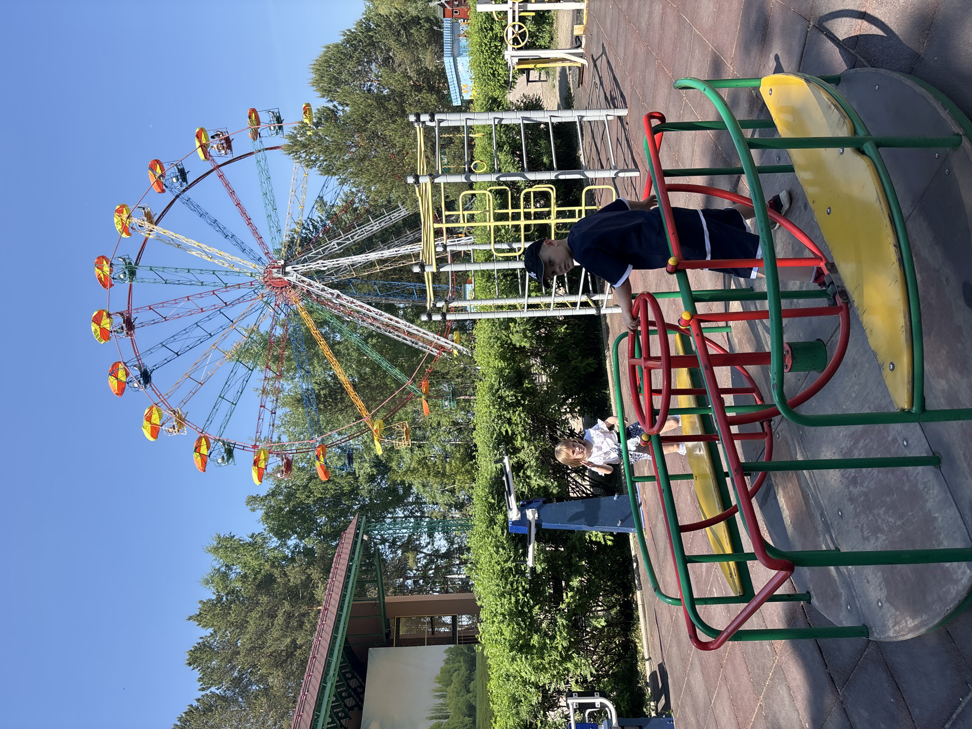 The image shows a playground in the city of Chaikovsky, Russia. A large, colorful Ferris wheel dominates the background, standing behind lush green trees. In the foreground, a boy in a dark blue shirt and cap stands on a playground merry-go-round structure made of green and red metal. A younger child in a white shirt is nearby, on a blue and silver piece of playground equipment, separated by a green hedge. The playground surface appears to be tiled. The sky is clear blue. A metal climbing structure is also visible in the mid-ground. A building is visible behind the hedge.