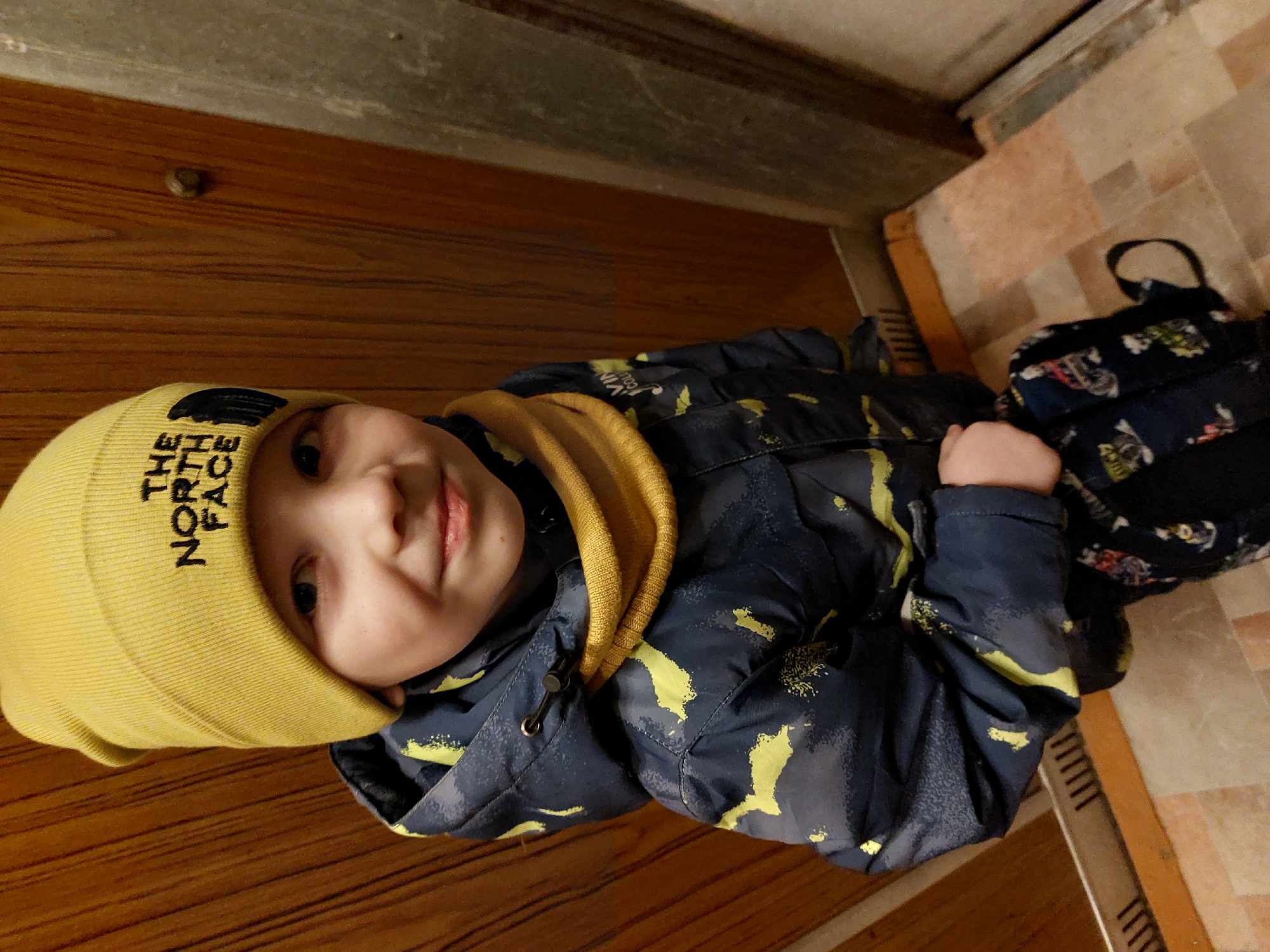 A small child wearing a yellow beanie with the words "The North Face" printed on it and a dark blue jacket with yellow patterns is lying on a wooden floor. The child has light skin and is smiling slightly. Next to the child, partially on a tiled floor, is a dark blue backpack with a floral pattern. The background includes parts of a wooden wall and a ceiling. The location is in a building in Chaikovsky, Russia.