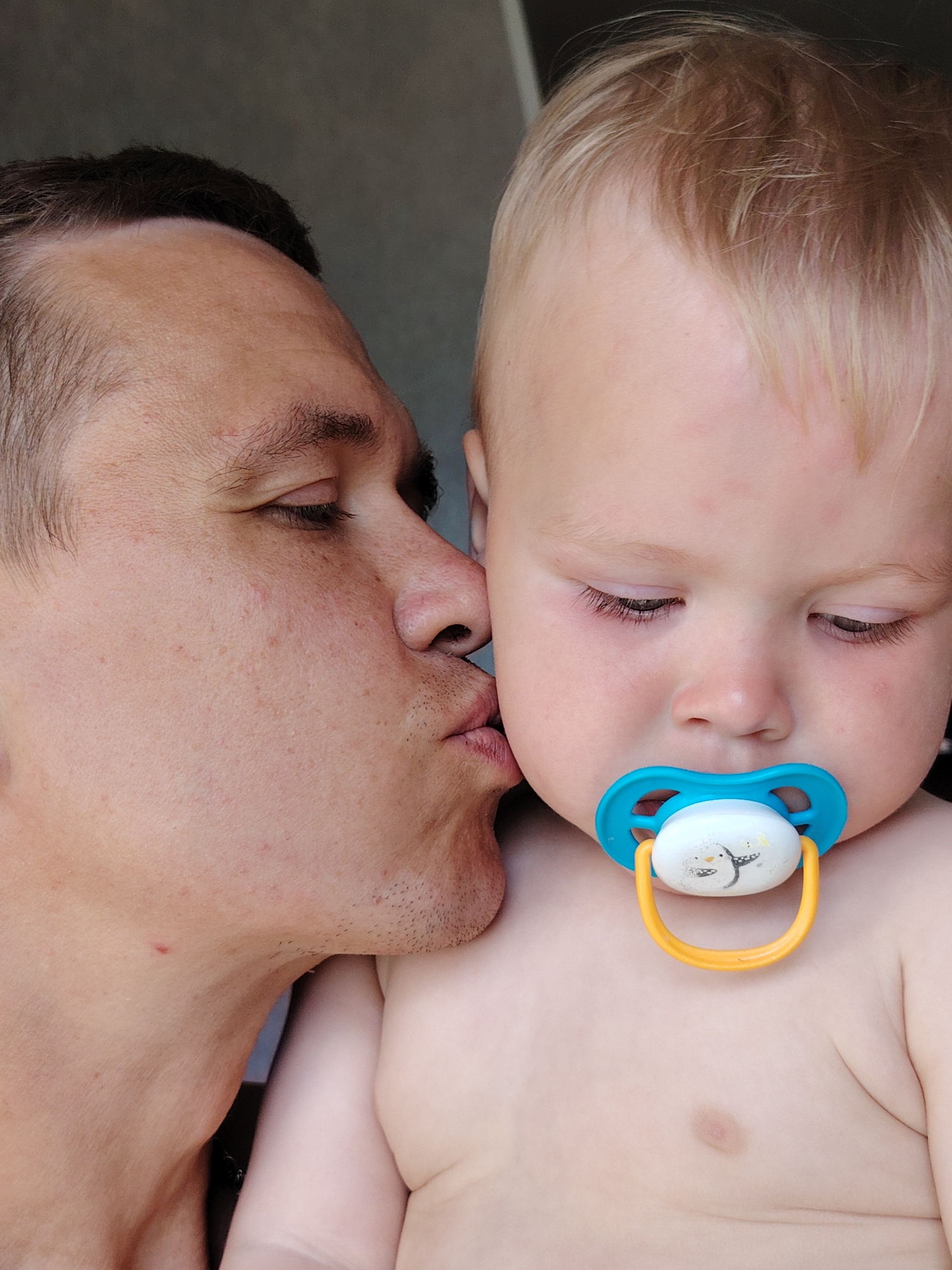 A close-up, portrait-oriented image shows a man kissing a baby. The man, positioned on the left side of the frame, is leaning in to kiss the baby's cheek. His eyes are closed, and his lips are pressed against the baby's skin. The baby, occupying the right side of the frame, has its eyes downcast and is wearing a blue pacifier. The background is a dark, plain surface. This image was captured in Chaikovsky, Russia.