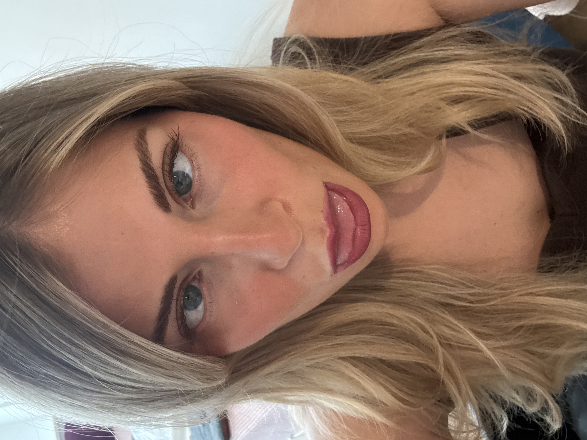 The image is a close-up portrait of a young woman, captured from a slightly high and tilted angle, suggesting a selfie. The woman has long, wavy blonde hair with visible darker roots, framing her face. She has fair skin, pronounced dark eyebrows, and light-colored eyes, appearing to be blue or green, which are looking directly at the viewer. Her face shows applied makeup, including eyeliner or mascara on her eyelashes, a touch of blush on her cheeks, and glossy, deep pink or berry-colored lipstick. Her mouth is slightly open, revealing a glimpse of her tongue and teeth, contributing to a playful or alluring expression. The background is largely out of focus, consisting of bright, light tones on the left and upper parts, possibly a wall or ceiling, with some darker, indistinct elements on the right. The lighting is soft and bright, suggesting an indoor setting during the daytime. No specific location cues are visible within the image to indicate Paris, France, but the image is provided in that context. There is no visible text in the image.