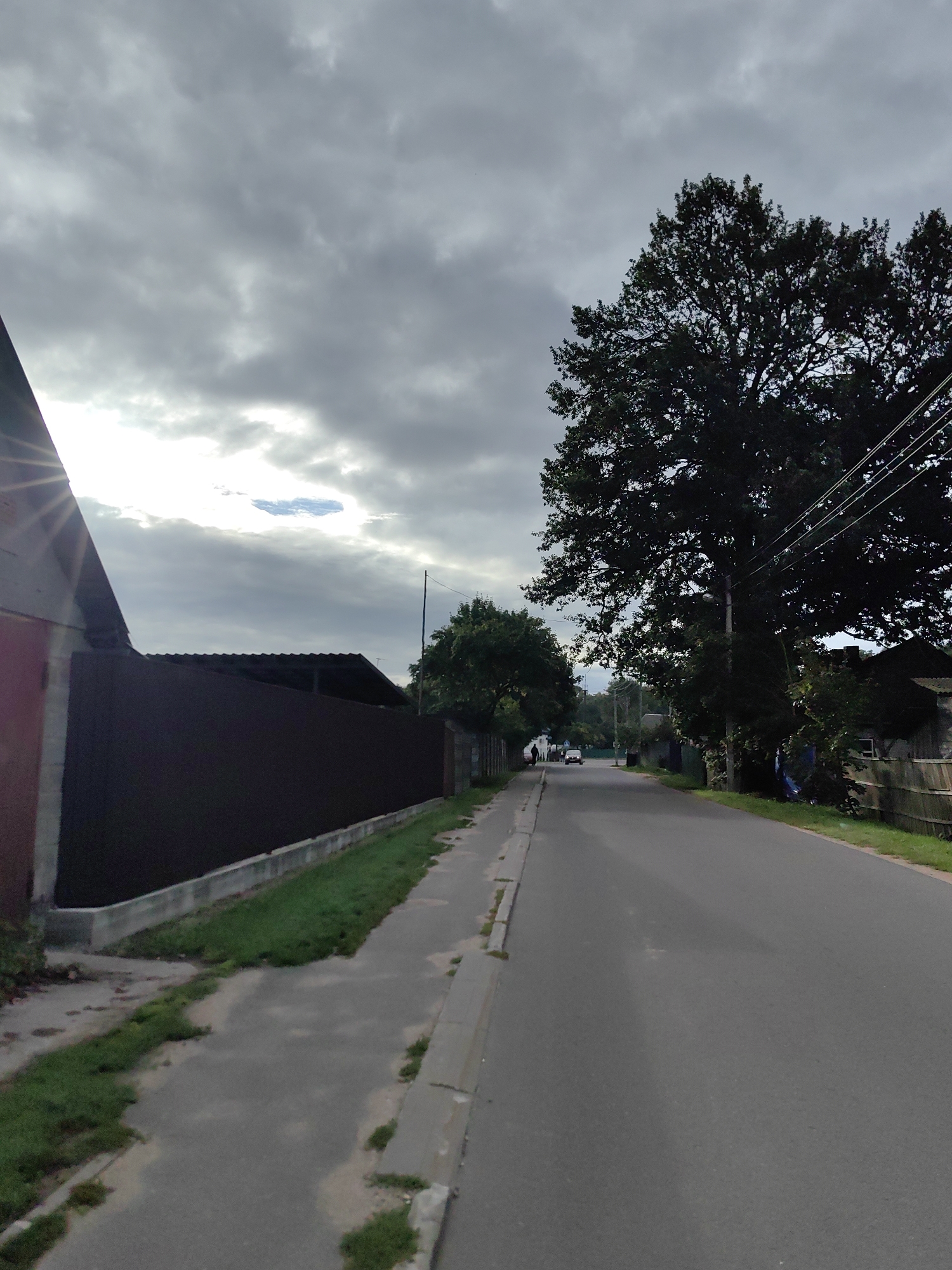 The image depicts a street scene in Fanipal, Belarus. On the left side of the frame, a dark brown metal fence runs along a paved walkway and a grassy verge. A building with a red brick facade and a grey roof is partially visible on the far left, with sunlight glinting off its surface. The sky is overcast with dramatic, dark clouds, though a patch of bright light breaks through. A large, dark green tree dominates the right side of the image, its branches reaching towards the sky and obscuring a telephone pole and power lines. The paved street extends into the distance, with a few vehicles visible. Houses and more trees line the right side of the street.
