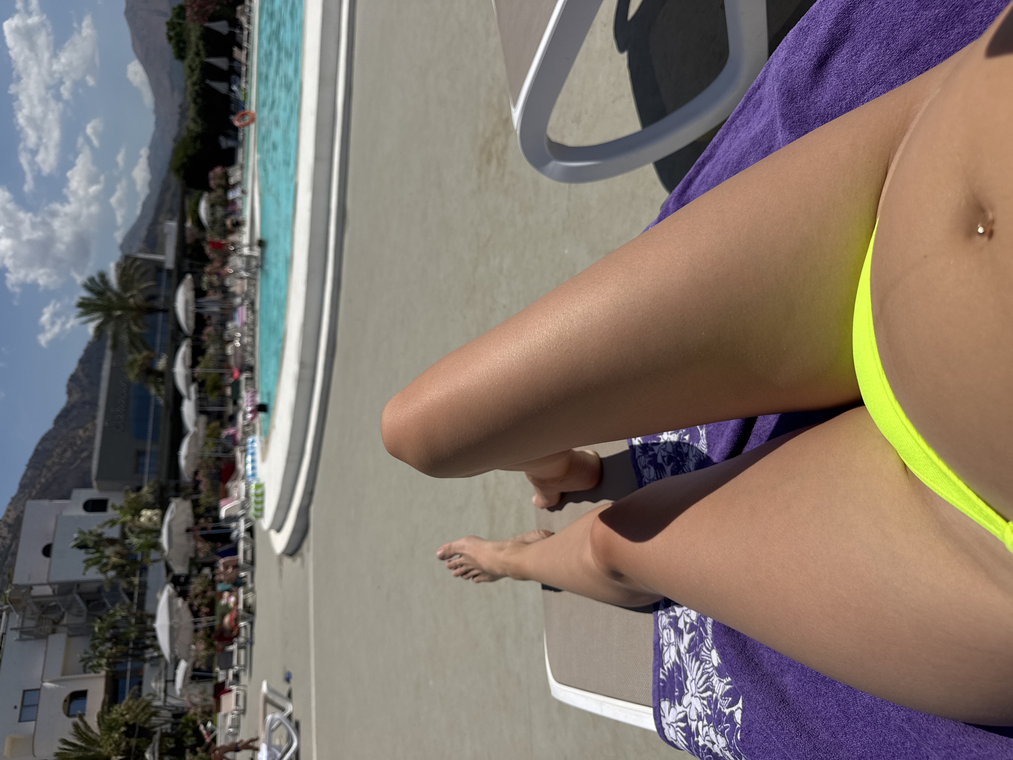 A first-person perspective shot shows a person relaxing on a lounge chair by a swimming pool on a sunny day. The person's legs are prominently displayed, clad in a neon yellow bikini. A purple towel with a white floral pattern lies beneath them. In the background, a resort-style building with white walls and balconies is visible, surrounded by palm trees and mountains under a partly cloudy sky. The swimming pool area features lounge chairs, umbrellas, and people enjoying the water and sun. The location is identified as Isola delle Femmine, Italy.