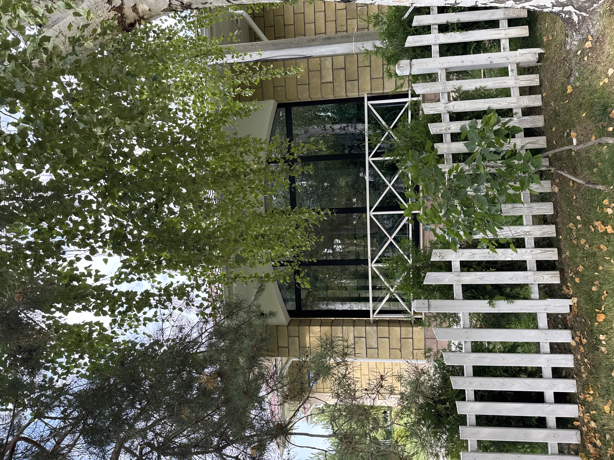 The image depicts the exterior of a house in Sary-Oy, Kyrgyzstan. The house features a light yellow brick facade and a large, dark-framed window. In front of the window is a white railing with a decorative criss-cross pattern. A white picket fence runs across the foreground of the image, partially obscuring a small sapling growing in front of it. The ground is covered in green grass and scattered fallen yellow leaves. Trees with lush green foliage overhang the scene, with some branches reaching down towards the house and fence. A portion of a white building with a red roof is visible to the left of the house. A pale grey utility pole stands to the right of the house. The sky is visible through the leaves of the trees.