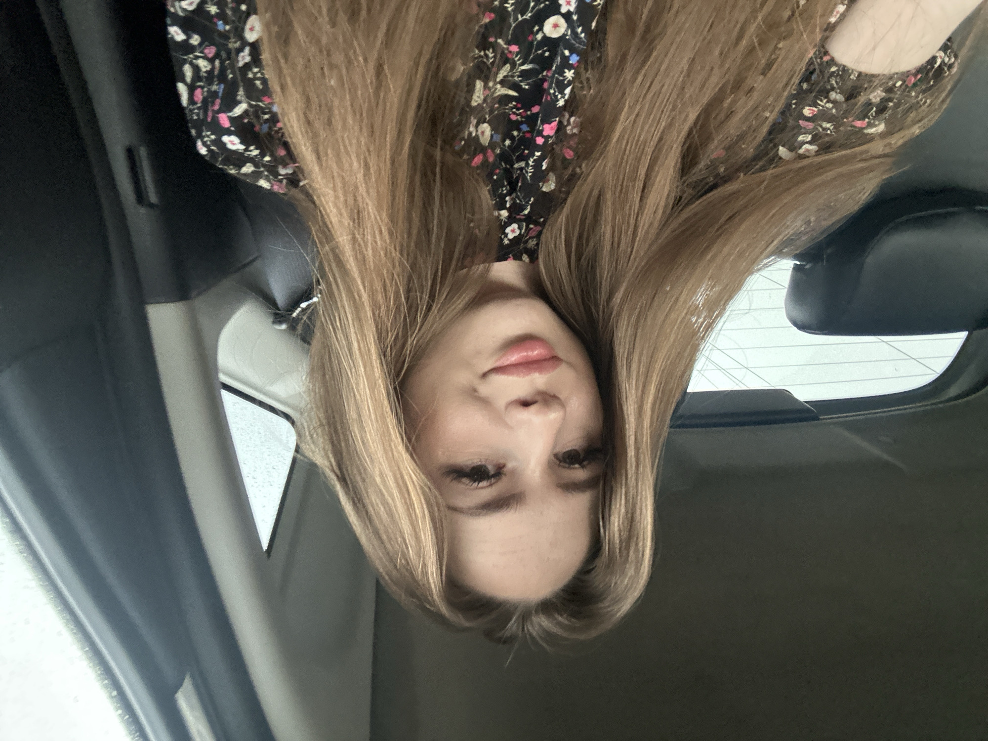A young woman with long, blonde hair is sitting in the passenger seat of a car. She is wearing a black dress with a floral pattern. Her hair is falling around her shoulders and partially obscuring the background, which appears to be the interior of a car. The woman is looking at the camera and smiling slightly. The image is taken from a slightly elevated angle, looking down at the woman. The lighting is natural, likely from the car's windows, and the overall mood is casual and relaxed. The location is unknown.