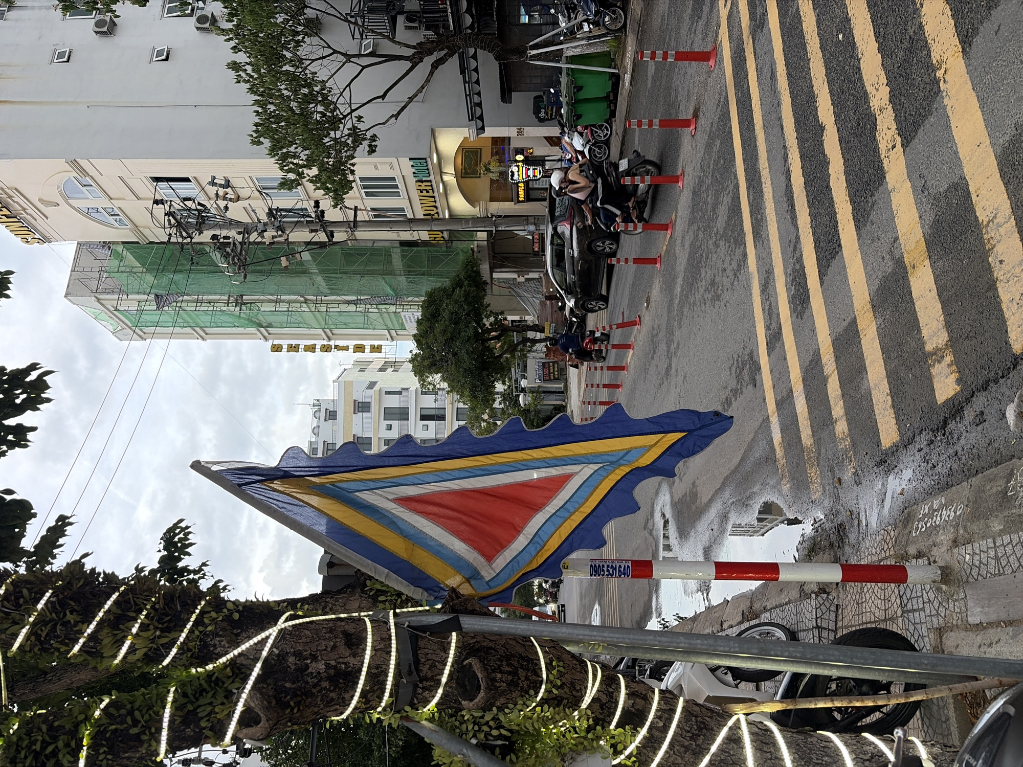 In an urban setting in Đà Nẵng, Vietnam, a flag with a triangular red center surrounded by blue, yellow, and white bands waves from a pole next to a tree wrapped with white string lights. The street is wet from recent rain, with puddles reflecting the overcast sky and surrounding buildings. A pedestrian crossing with yellow stripes is visible on the road. Several motorcycles and scooters are parked on the side of the street, and a few are in motion on the road. Two people ride on one scooter. Buildings line the street, including a hotel with a sign that reads "SUNFLOWER HOTEL". Construction is visible on one of the buildings, with green netting covering scaffolding. The scene captures a typical day in a Vietnamese city, with a mix of traffic, pedestrians, and urban infrastructure.