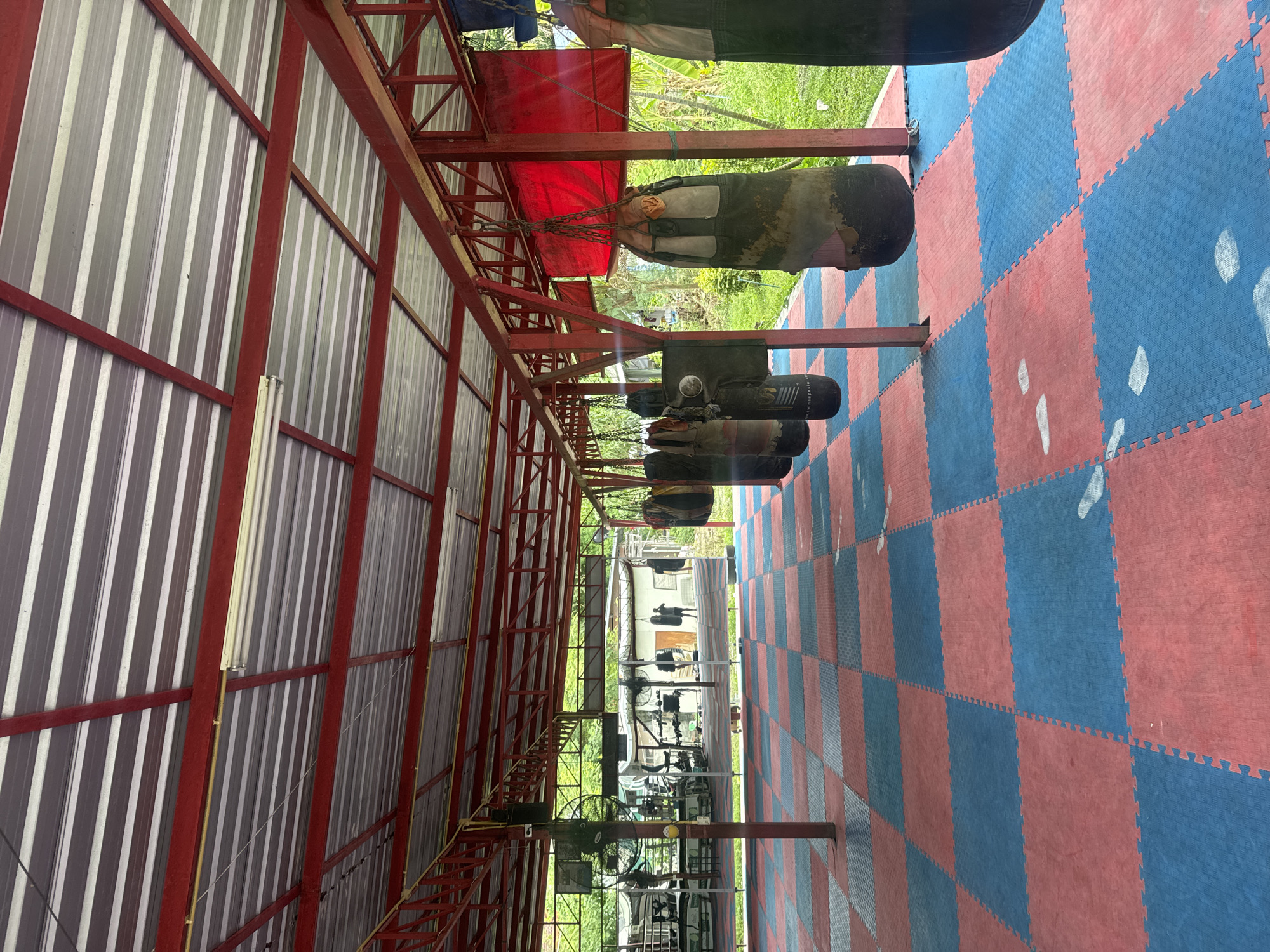 The image depicts the interior of a gym in Thong Sala, Thailand. The structure has a red steel frame, with a grey corrugated metal roof and walls. Hanging from the frame are several punching bags. The floor is covered with interlocking red and blue foam mats. There is exercise equipment in the background of the scene. The scene is in daylight.