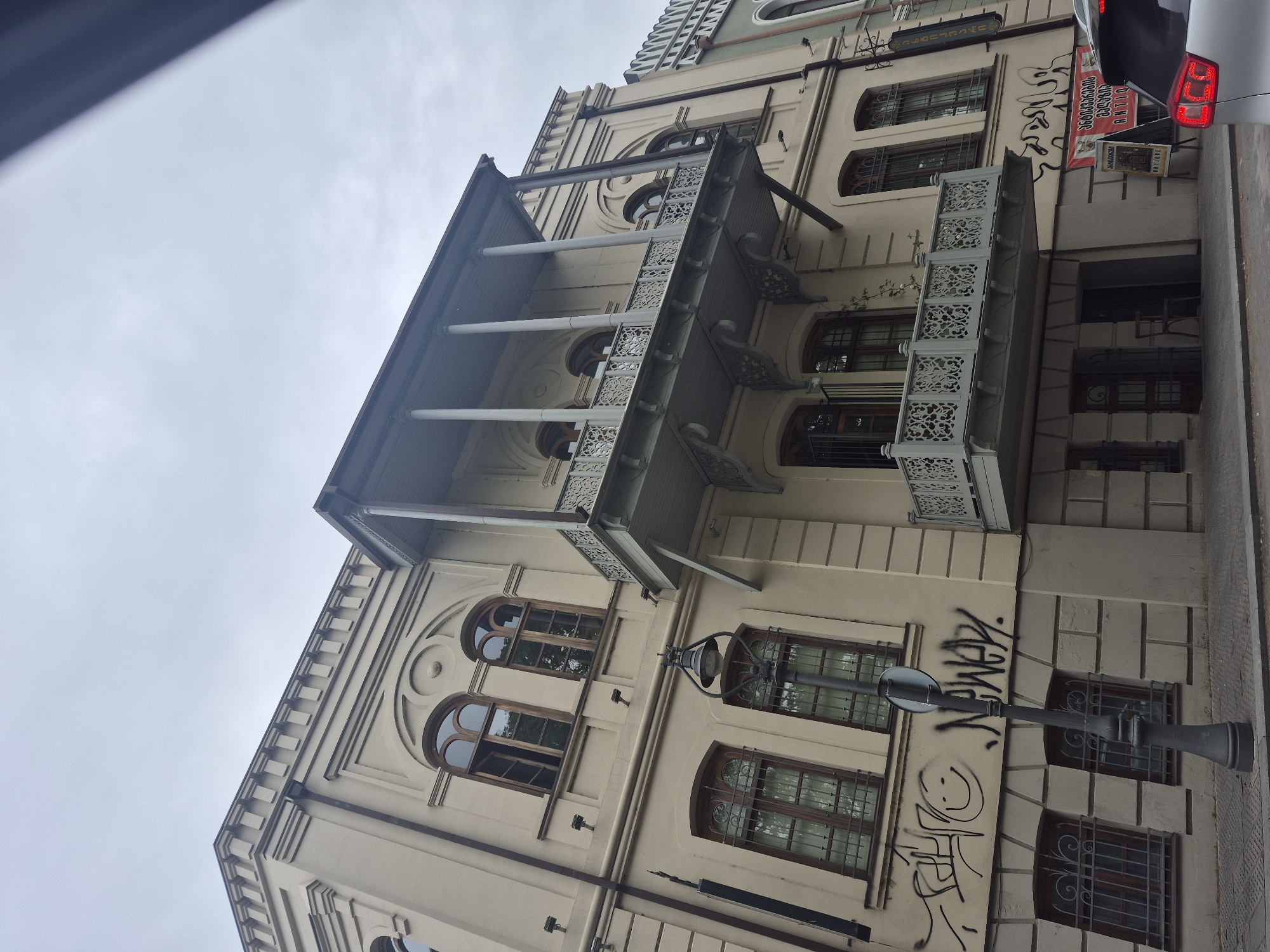 The image showcases a multi-story building in Tbilisi, Georgia, characterized by its light-colored facade, arched windows with decorative grilles, and ornate balconies supported by columns. Graffiti is visible on the lower portion of the building's exterior. A street lamp extends from the building's side. A car and signage with Georgian text are visible on the right side of the frame. The sky is overcast.
