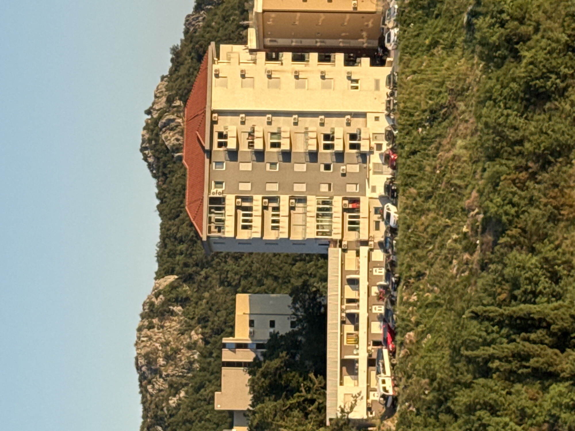 The image depicts a view of buildings and a hillside in Rafailovići, Montenegro. In the foreground, a steep, densely vegetated hillside is visible. Above this, a row of buildings is situated, with a low-rise structure in the center and taller apartment buildings on either side. The central low-rise building has a colonnaded walkway or balcony area facing the viewer. A line of parked cars is visible along the front of these buildings, including a white truck or van, a red car, and several other passenger vehicles. Behind this row, taller apartment buildings dominate the scene. The building in the center-right is a multi-story structure with a red tiled roof and numerous balconies equipped with air conditioning units. To its right, a portion of another taller, lighter-colored building is visible. In the background, a rugged, tree-covered mountainside rises under a clear blue sky. The lighting suggests it is late afternoon or early evening, with the sun casting a warm glow on the buildings.