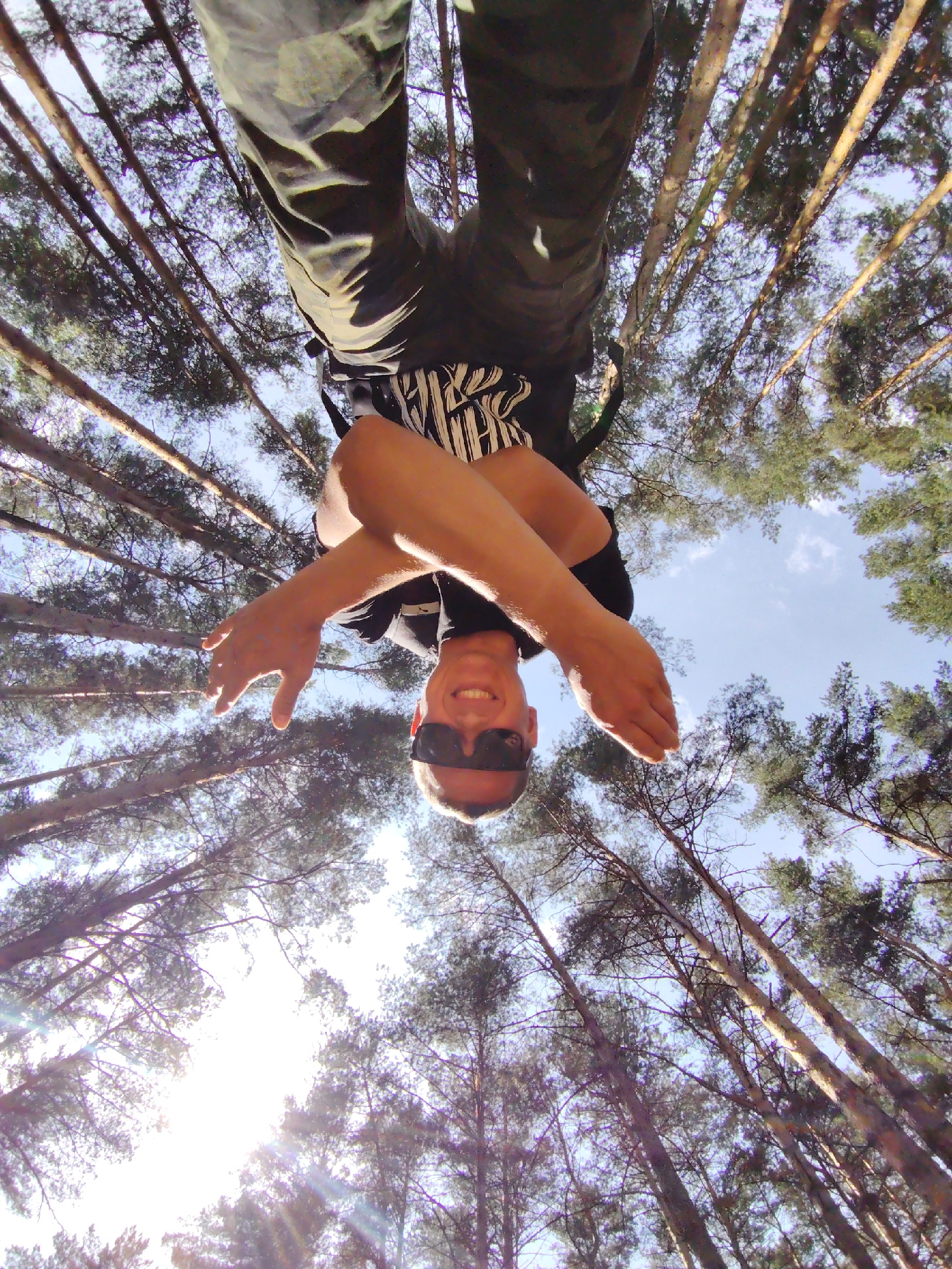 A man is inverted and facing the camera, likely suspended in the air, with his arms crossed and wearing sunglasses. He is in a forest, evidenced by numerous tall trees with dense foliage reaching toward the sky. Sunlight is filtering through the branches. The location appears to be in Chaikovsky, Russia, based on the context.