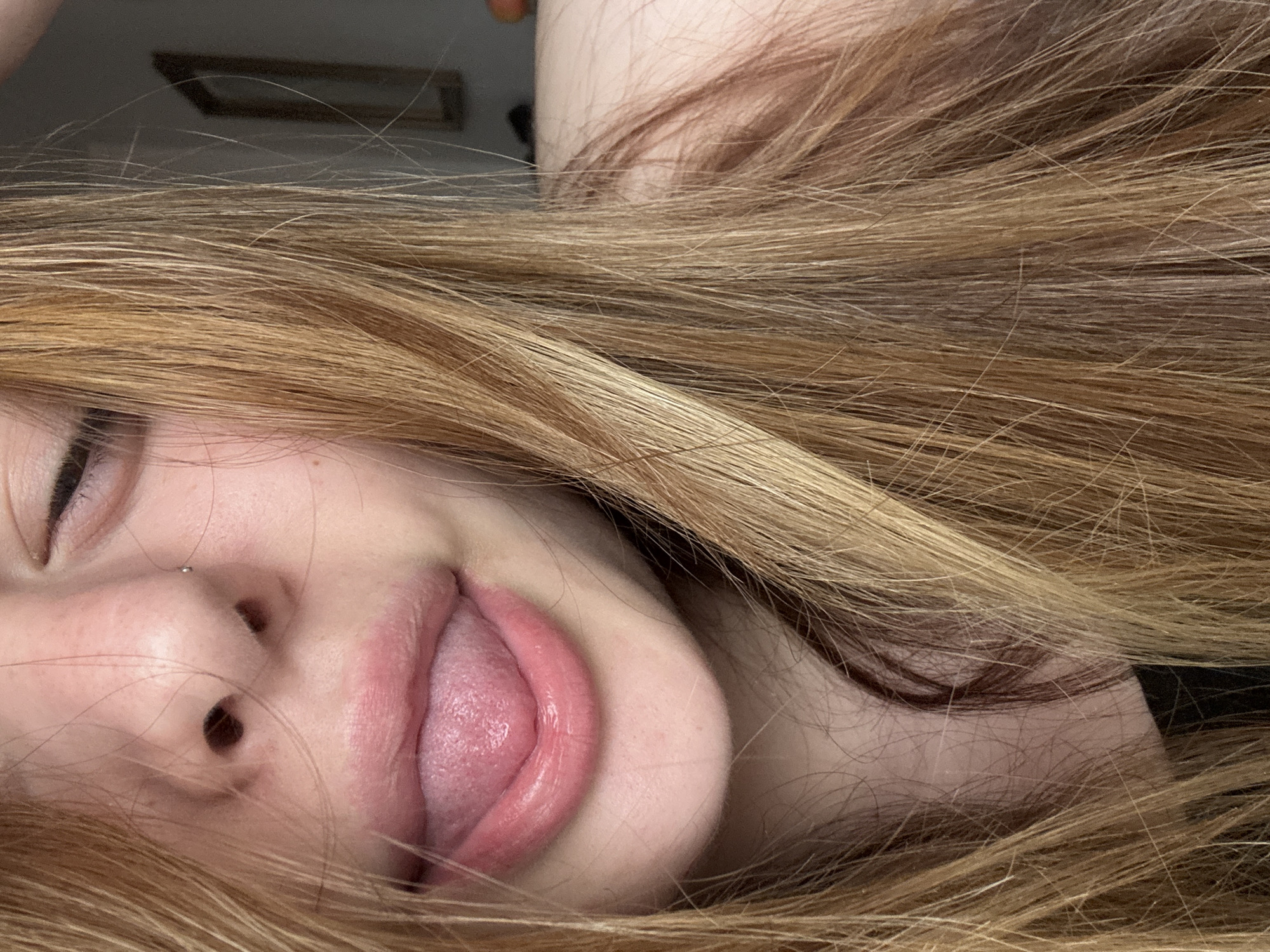 The image is a close-up, portrait-style shot of a person's head, oriented horizontally with the person's face on the left and hair extending to the right. The primary subject is a person with long, light brown and blonde hair, which dominates much of the frame, appearing to flow around their face.
The person's face is shown in profile or partial profile, with their left eye mostly closed or squinting. A small, silver stud nose piercing is visible on their right nostril. Their mouth is open, with their tongue extended outwards, suggesting a playful or expressive gesture. The lips are a natural pink hue. The skin appears fair with some freckles visible.
In the blurred background, through strands of hair, a dimly lit indoor setting is implied, with a dark, rectangular object, possibly a picture frame, partially visible against a lighter wall. There are no clear indications of the time of day or weather. No visible text is present in the image. The location context provided is Troyan, Bulgaria, although this is not discernible from the visual information alone.