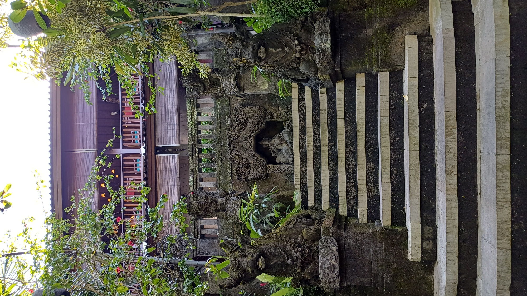 The image captures a stone stairway, potentially in a garden or temple setting in an unknown location. The stairs are constructed from alternating light and dark-colored stone steps, seemingly aged and worn. Adjacent to the stairs, there are intricately carved stone structures adorned with figures and decorative elements, possibly deities or mythical creatures. Vegetation, including green plants and flowering bushes, frames the scene. In the background, a multi-tiered wooden building with latticework is partially visible, suggesting a traditional architectural style. The image is oriented vertically.