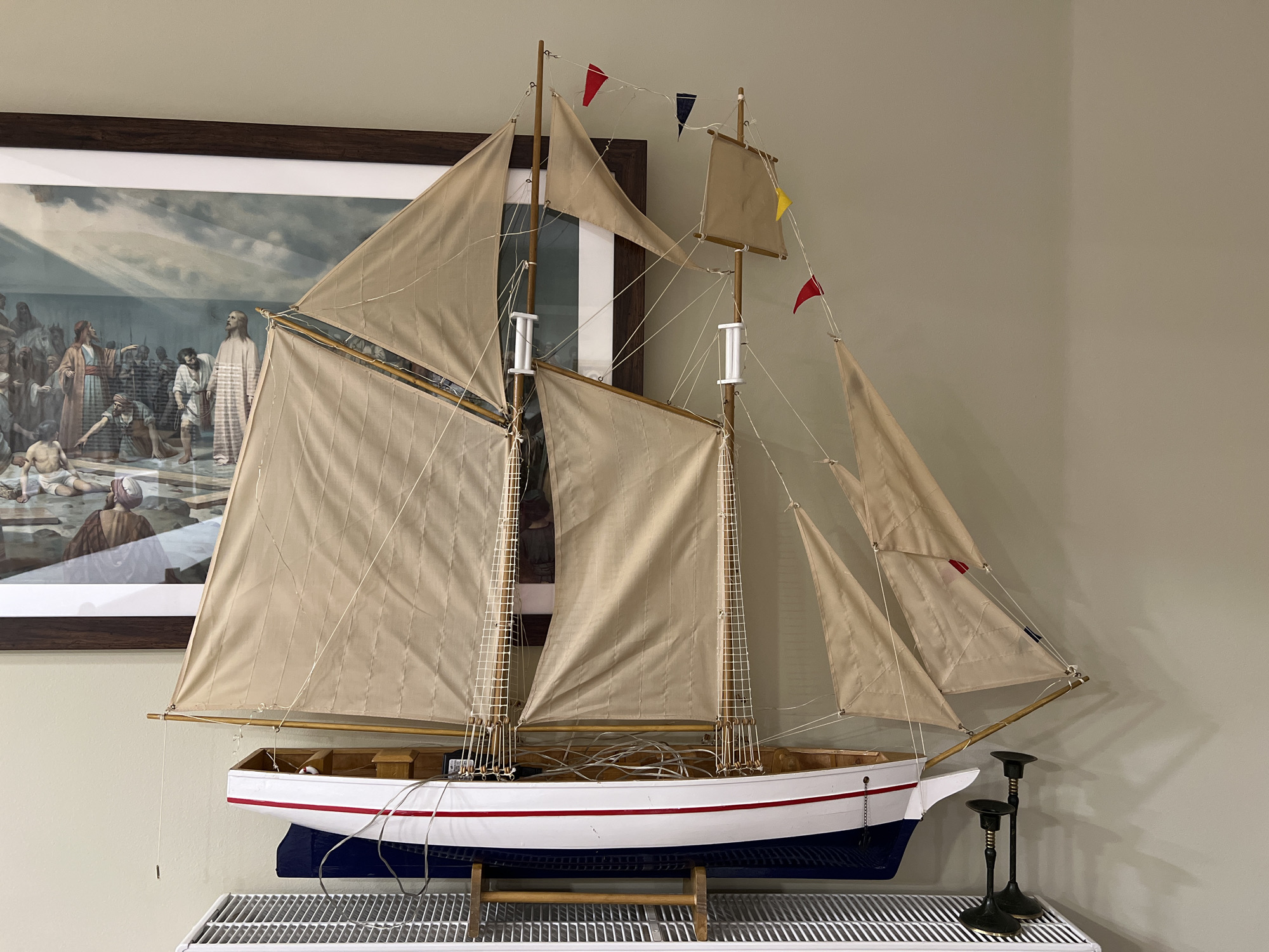 A model sailboat with tan sails is displayed on a white radiator cover, which is positioned in front of a light green wall. Above the sailboat, a framed picture depicting a scene with multiple figures in historical attire is hung on the wall. The sailboat is white with a red stripe along its hull and rests on a dark blue stand. The image shows a close-up of the sailboat's rigging, masts, and sails, as well as the framed artwork. Two black candlesticks with gold accents are placed to the right of the sailboat. The overall impression is a still life arrangement. The location is identified as Kallithea, Greece, however, there are no visual cues within the image to confirm this location.
