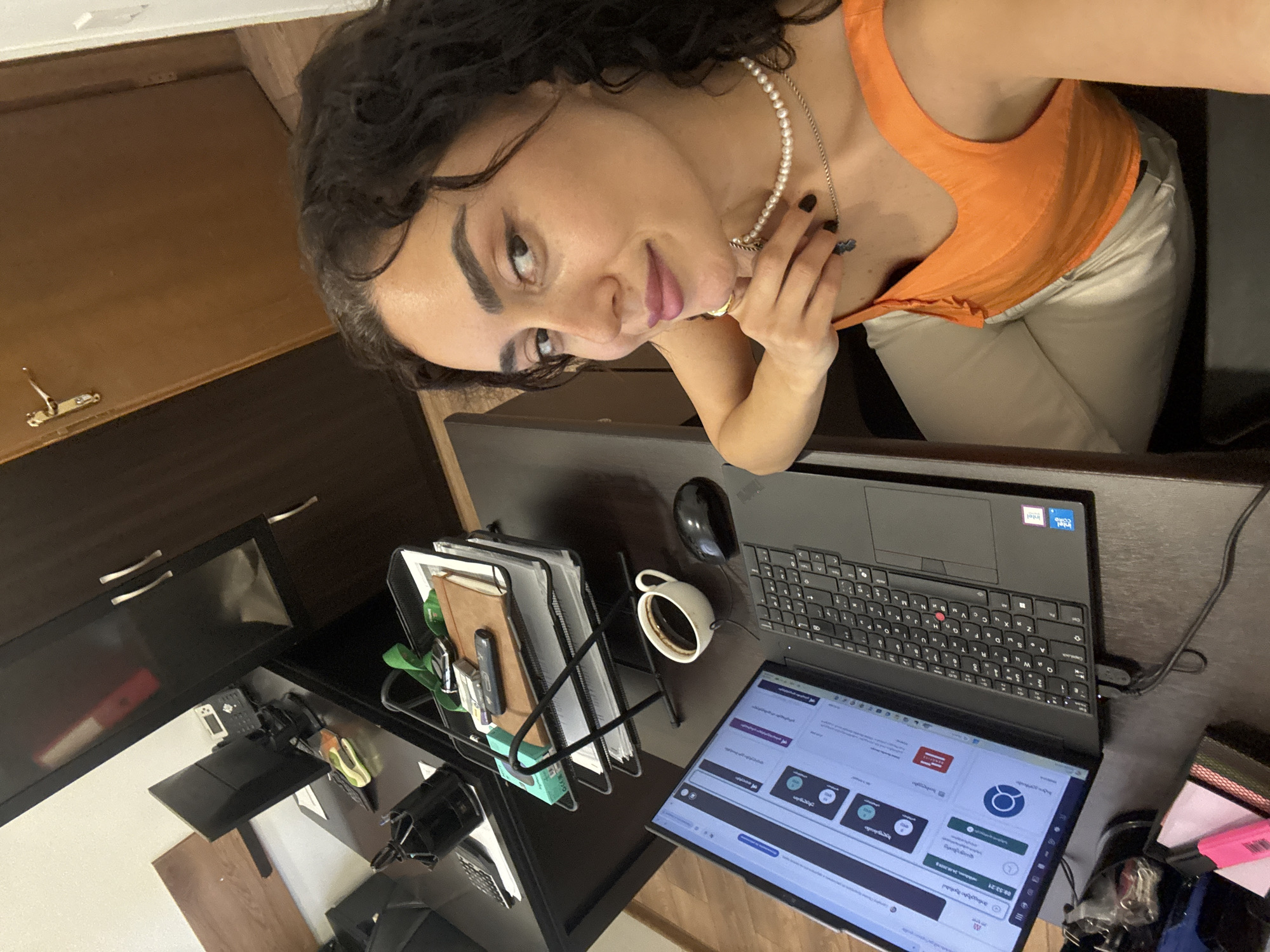 A woman with curly dark hair is taking a selfie in an office. She is wearing an orange top and light-colored pants, with a pearl necklace. She is seated at a desk with a laptop, a cup of coffee, and a mouse. In the background, there are office cabinets and a monitor. The environment appears to be an office in the city of Tbilisi, Georgia.
