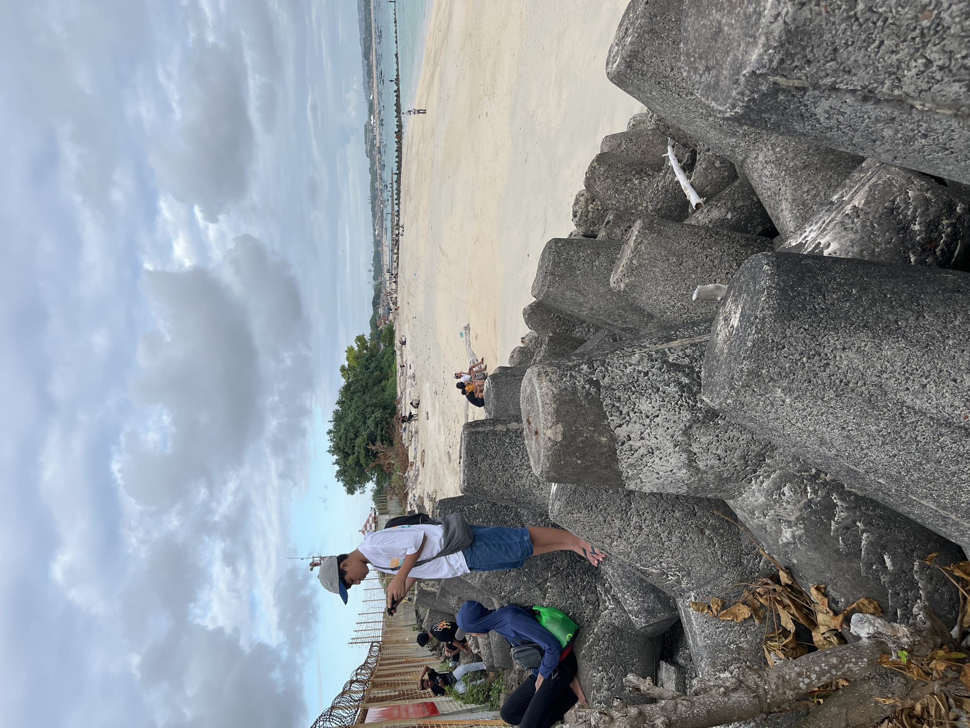 In Kuta, Indonesia, on a cloudy day, a group of people are gathered on a sandy beach near a breakwater made of large concrete tetrapods. In the foreground, two young males are seated on the tetrapods. One, wearing a blue hoodie and black pants, has a green bag on his lap. The other, with dark hair and a white t-shirt, sits with his legs crossed. A third male, wearing a white t-shirt, blue shorts, and a grey baseball cap, stands on a tetrapod, looking at his phone. He has a grey bag slung over his shoulder. Behind them, a fence with barbed wire runs along the top of the breakwater. Further down the beach, other individuals are visible sitting and walking on the sand. The ocean stretches out to the horizon, with boats anchored in the distance. The sky is filled with overcast, grey clouds. The environment appears to be a public beach area, possibly a tourist destination.