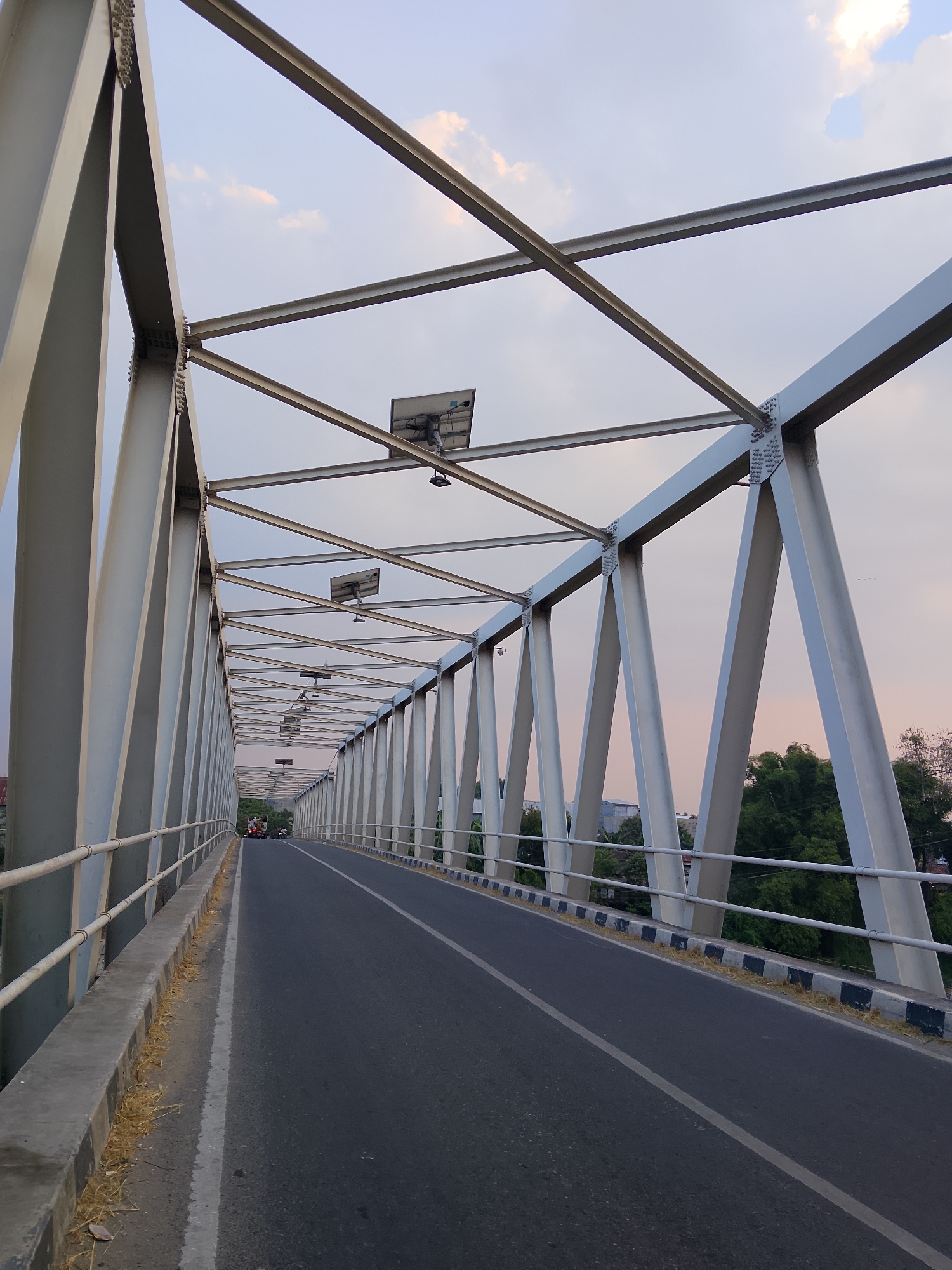 The image shows a view from a pedestrian walkway looking down a long, steel truss bridge. The bridge has a white metal structure with diagonal beams supporting a road. The road is dark asphalt with a white lane divider. On the left side of the road, there is a concrete walkway with a white metal railing. There are solar panels mounted on top of the bridge's structure. In the distance, several motorbikes are visible on the road, and the bridge is lined with trees and some buildings on the right side. The sky is partly cloudy and has a pale pinkish hue, suggesting it is either sunrise or sunset. The location is identified as Betet, Indonesia.