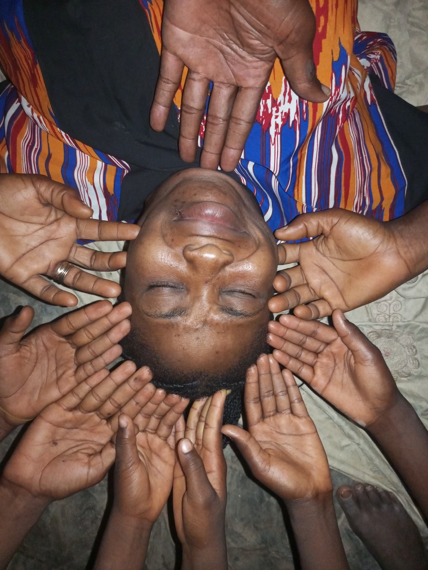 The image depicts a person lying down, possibly on a bed, with their eyes closed. Their head is positioned in the center of the frame, and multiple hands are surrounding their face. The person is wearing a black garment underneath a vibrantly patterned cloth with stripes of orange, blue, red, and white. The hands appear to be of different sizes and are in various positions, some touching the person's face and others resting around it. The setting appears to be indoors, with a textured blanket or sheet visible beneath the person. The lighting suggests it may be nighttime or a dimly lit indoor space. There is no visible indication of the location being in the city of Tempure, Nigeria.