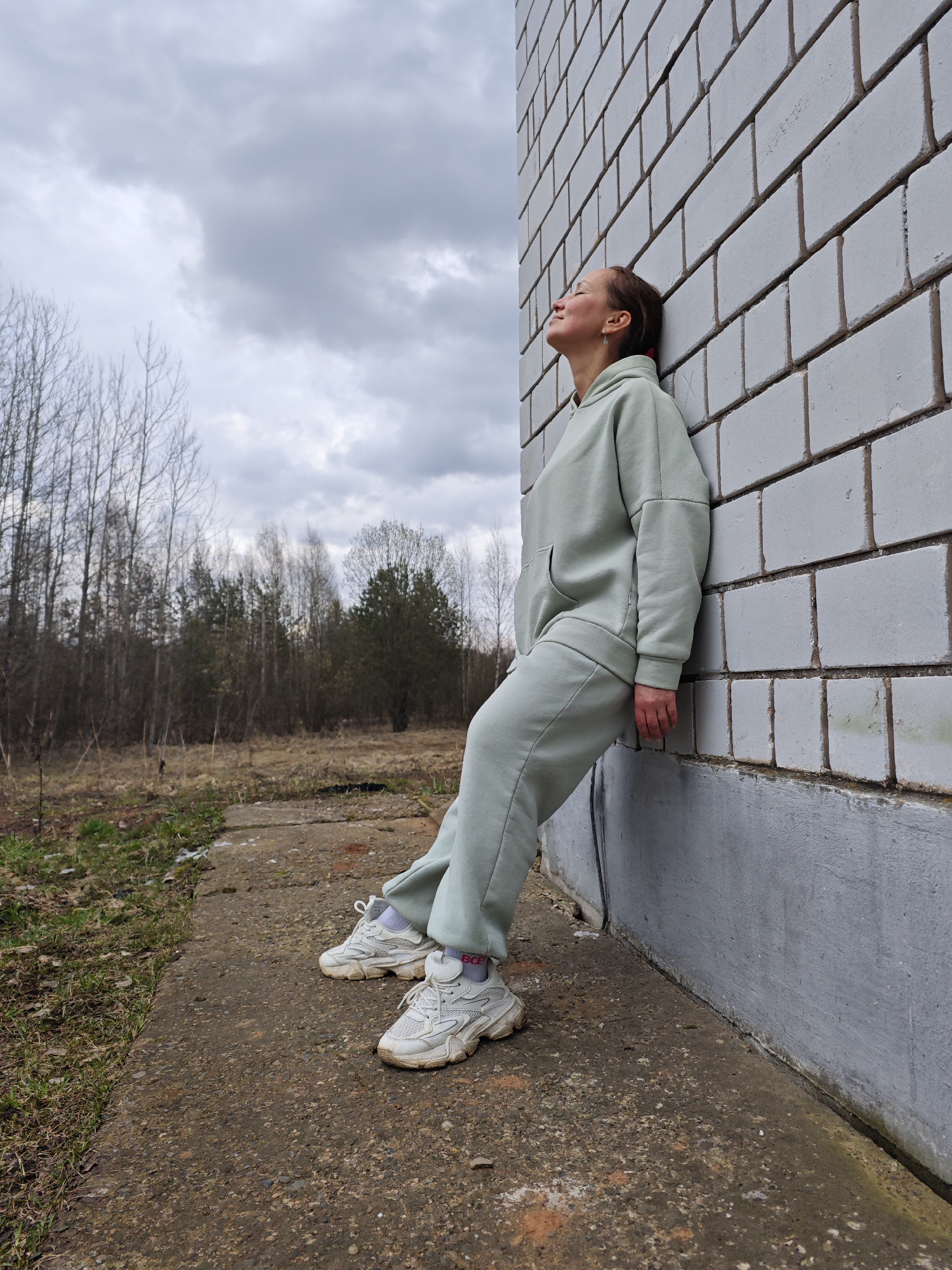 A woman is standing outside a brick building in Марковский, Russia. She is leaning against a white brick wall with her eyes closed and her face tilted up towards the cloudy sky. She is wearing a light green tracksuit and white sneakers. The ground is concrete, and there is a grassy field and bare trees in the background.