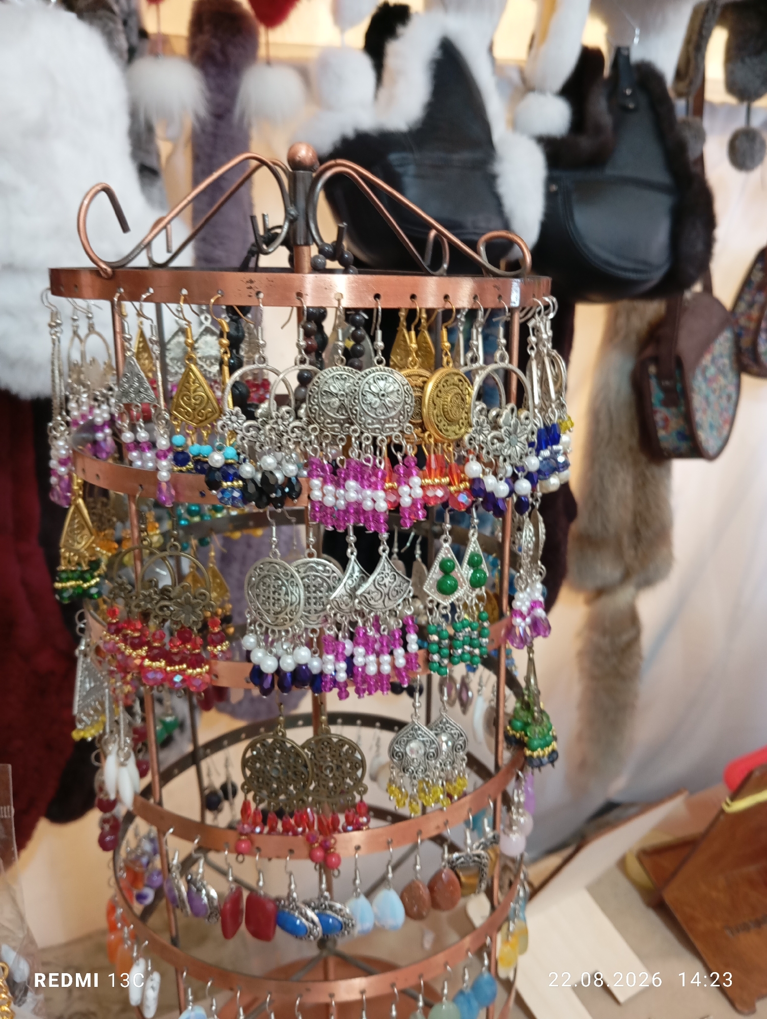 A multi-tiered copper earring stand displays a variety of colorful, ornate earrings. The earrings feature intricate metalwork, beads in shades of pink, purple, blue, green, red, and yellow, and some have dangling charms. The stand is positioned in front of a display of what appear to be winter hats and fur accessories, possibly in an outdoor market setting. The background is blurred, but suggests a retail environment. The bottom right corner of the image contains a watermark with the text "REDMI 13C" and a date and time stamp "22.08.2026 14:23". The location is identified as Amirsay, Uzbekistan.