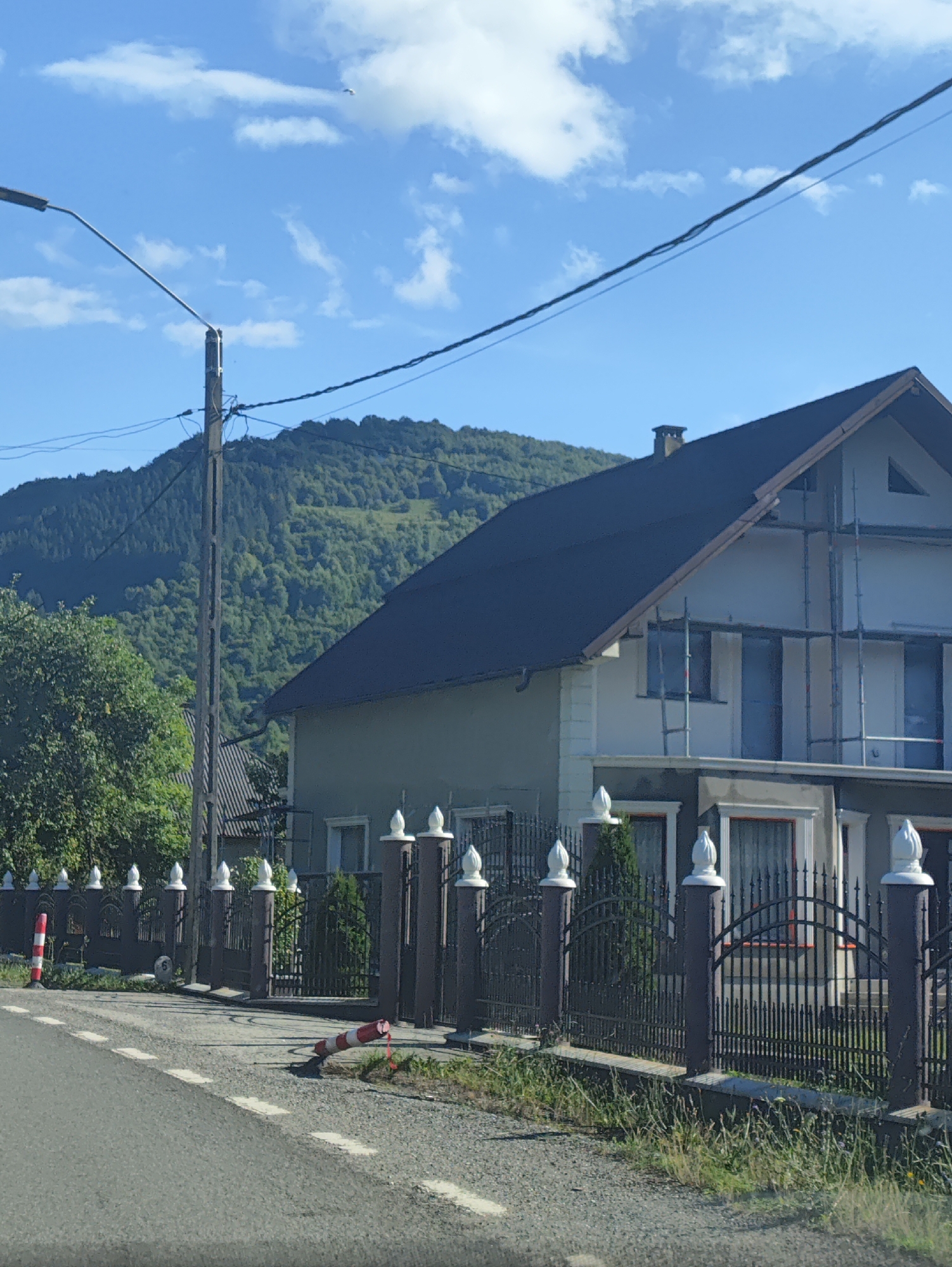 A street-level, eye-level, medium shot captures a residential property in Leordina, Romania, on a clear, sunny day. The scene is dominated by a two-story house, partially covered by scaffolding, suggesting ongoing construction or renovation. The house features a dark-pitched roof and light-colored exterior walls. A dark, ornate metal fence runs along the property line, with decorative finials atop its posts.
In the foreground, a paved road with white dashed lines separates the viewer from the property. A red and white striped traffic cone lies on its side on the shoulder of the road. Beyond the fence, several evergreen trees are visible, along with a backdrop of a densely forested hillside under a bright blue sky dotted with white clouds. A tall utility pole stands near the property, with electrical wires extending across the frame. The overall impression is of a quiet, rural residential area in Romania.