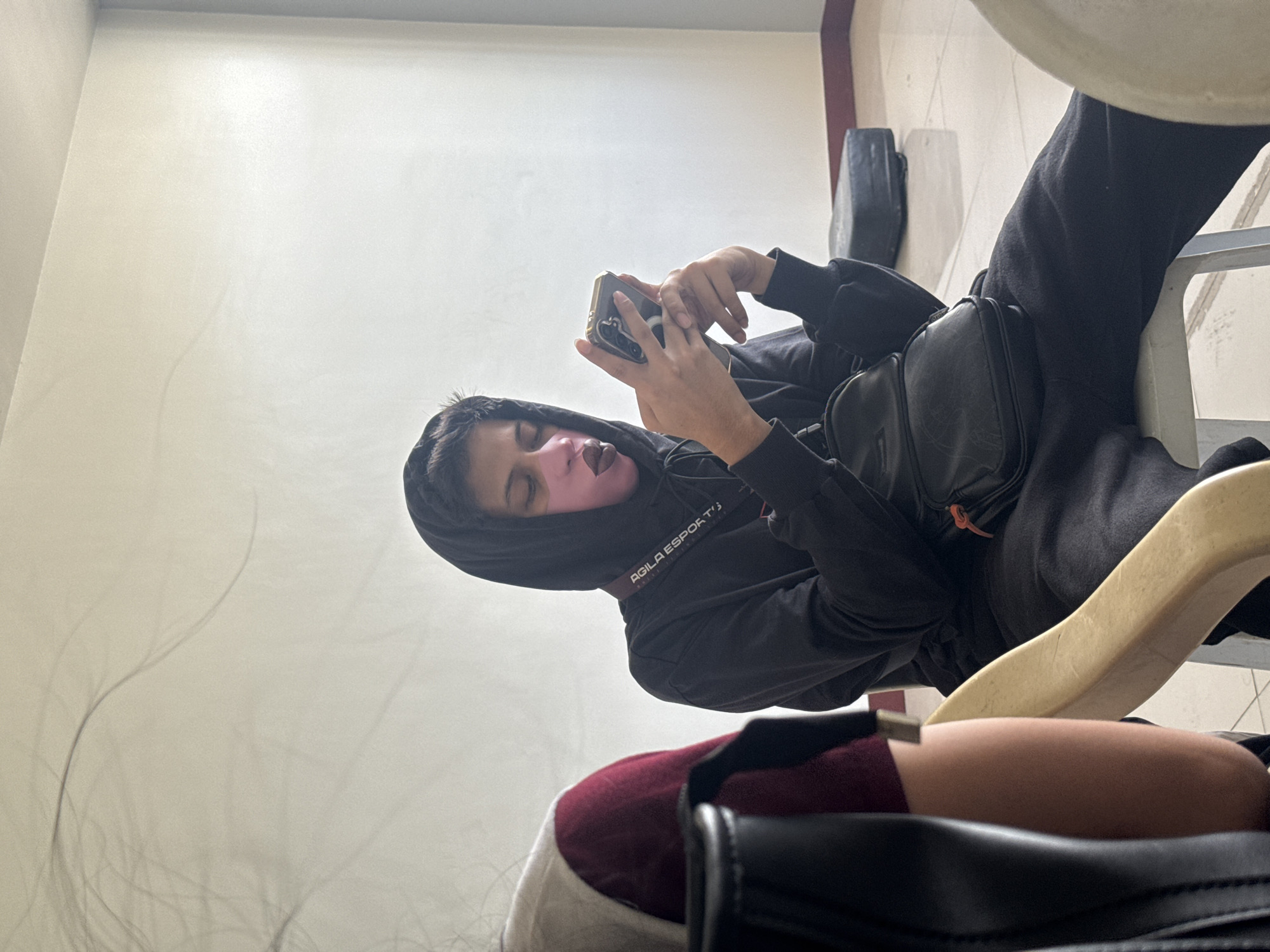 A person wearing a black hoodie and black pants is seated on a light-colored plastic chair, appearing to be using a mobile phone. The person has a black shoulder bag slung across their body. The scene includes a portion of a white wall and what appears to be another person's leg in the foreground. The location is Davao City, Philippines.