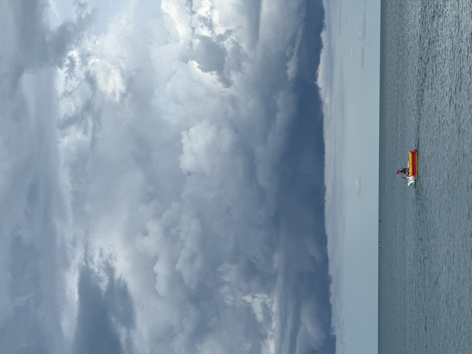 A single orange and white boat with a person aboard navigates the calm, gray waters of the ocean under a heavy, overcast sky. The horizon is a sharp line dividing the sea from the voluminous, dark gray clouds. In the distance, several other small objects, possibly boats or buoys, are scattered across the water. The scene appears to be located in the vicinity of Baan Chaweng Noi, Thailand, characterized by the expansive ocean and dramatic cloud formations.