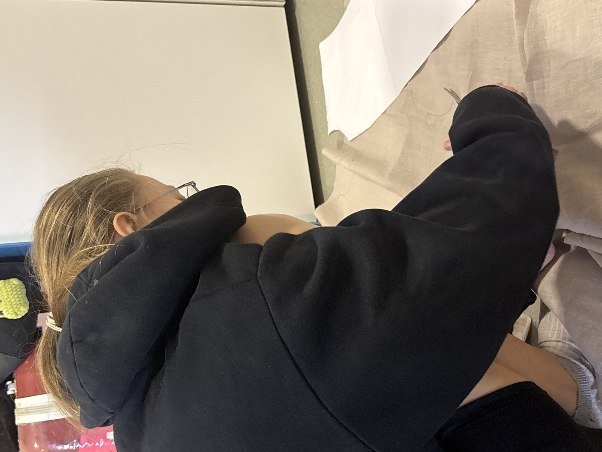 A young person with light-blonde hair and wearing glasses is hunched over a beige fabric on a surface. They are wearing a black hoodie and black pants. They appear to be cutting the fabric with scissors. On the fabric is a white paper pattern. To the left of the person is a rolling cart with objects on it. Behind the person is a white board.
The location of this photo is unknown and cannot be determined to be in Perm, Russia.