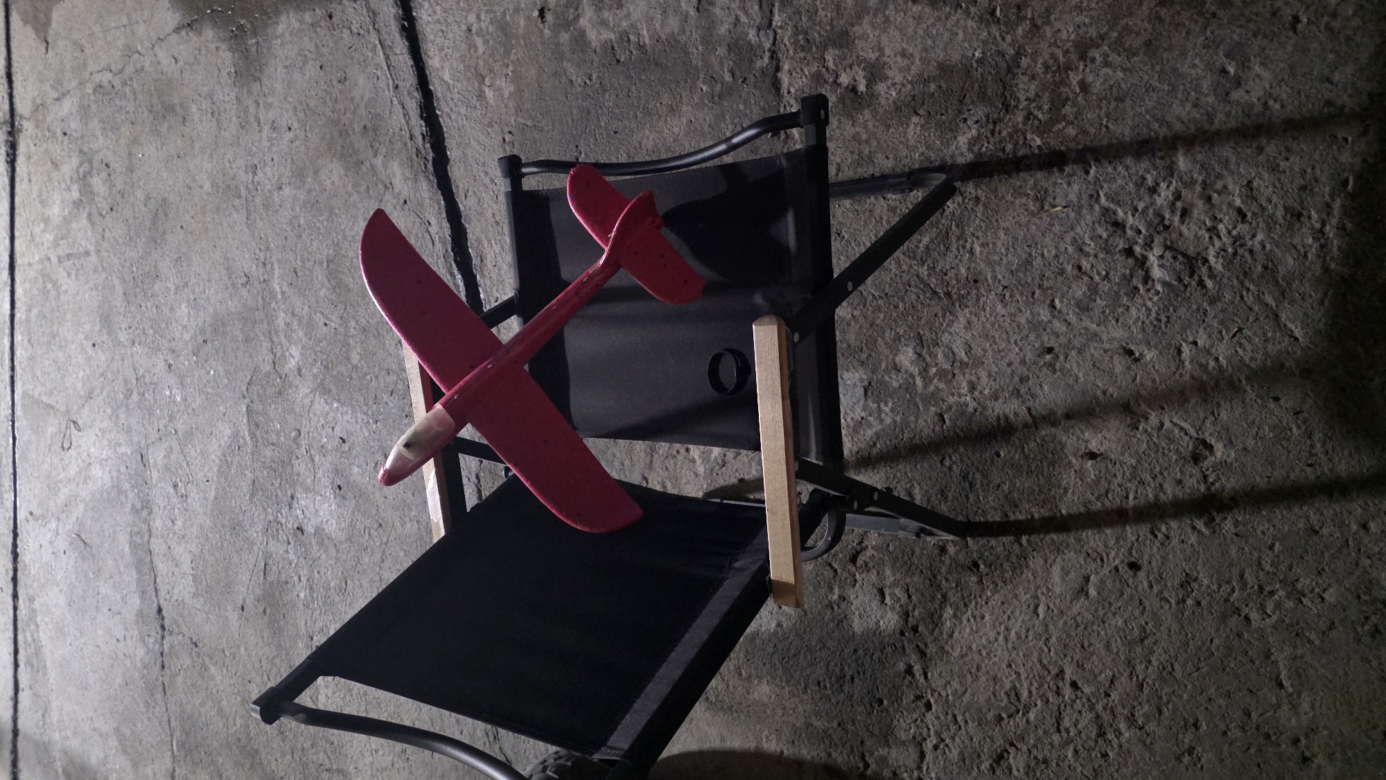 A close-up, overhead shot shows a red toy airplane placed on a black fabric camping chair with wooden armrests. The chair is situated on a rough, gray concrete surface. The scene appears to be outdoors in Chuy, Kyrgyzstan, possibly in an industrial or unfinished area due to the raw concrete and overhead lighting casting shadows. The airplane is positioned diagonally across the seat of the chair, with its nose pointing towards the upper left of the frame. The concrete exhibits cracks and uneven textures, characteristic of a weathered urban environment. The lighting suggests it is either dusk or nighttime, with artificial light sources creating defined shadows of the chair and the airplane on the concrete.
