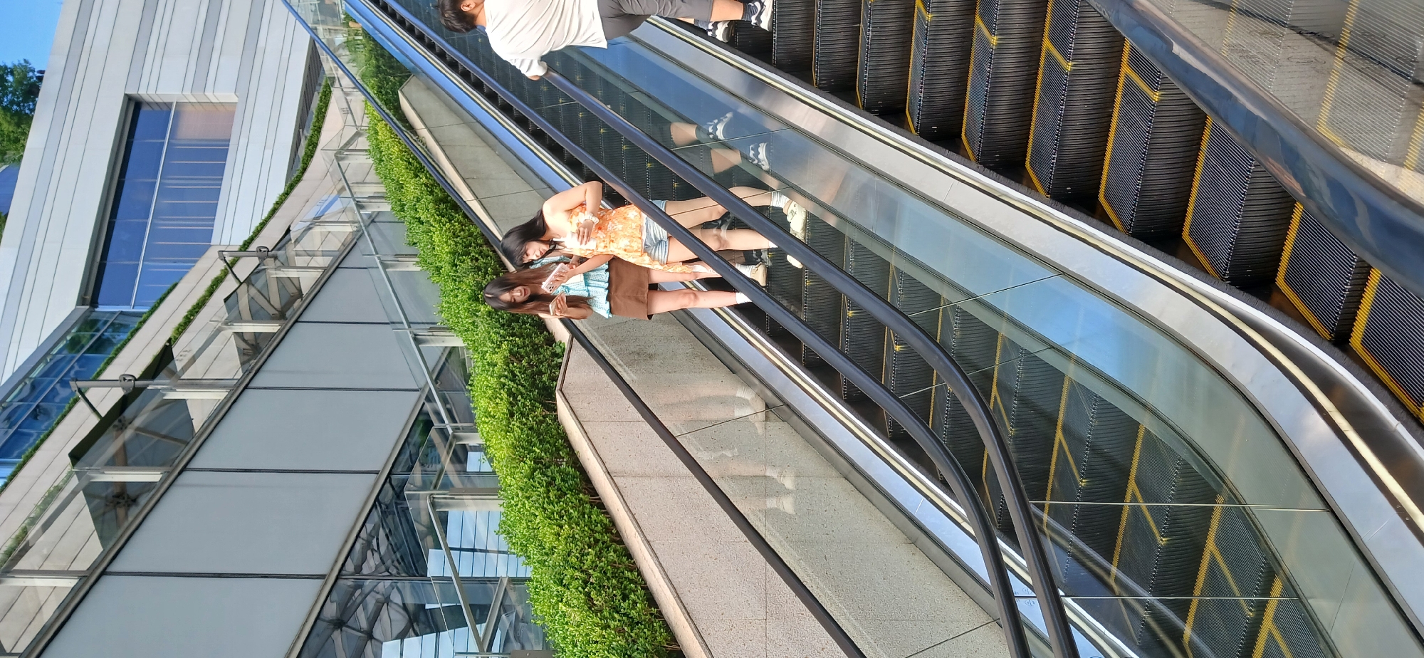 Two young women stand on an escalator in the Jiulongpo District, China, each holding a mobile phone and appearing to be in conversation. The woman on the left wears a light blue patterned halter top and a brown skirt, while the woman on the right sports an orange tube top and denim shorts. The escalator is flanked by a modern building with glass and metal accents, and lush green shrubbery. A man in a white t-shirt and grey shorts stands further up the escalator on the right. The escalator steps are black with yellow markings. The scene captures a moment of everyday life in an urban environment.