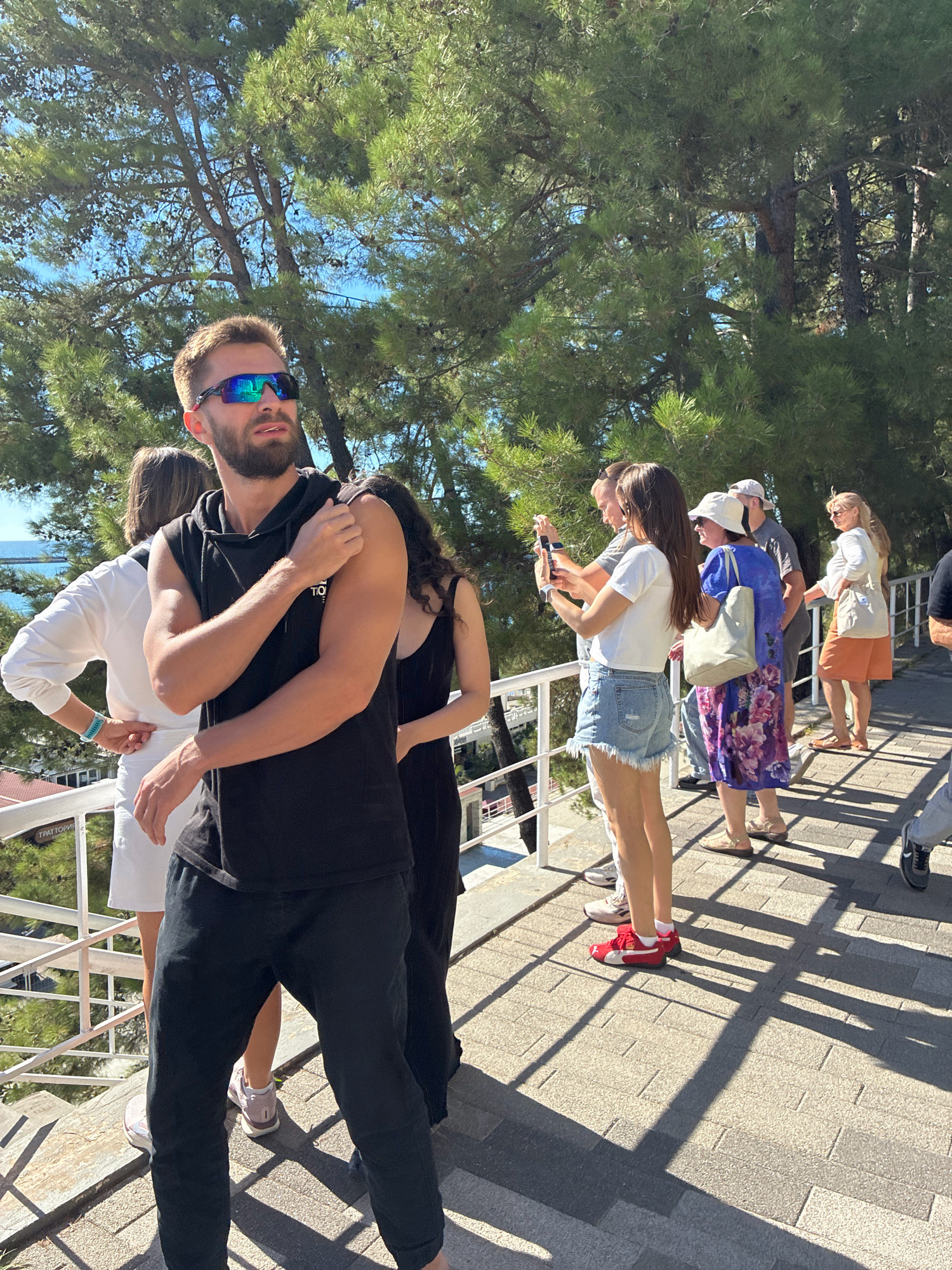 This is a photograph of a group of people on a sunny day, likely in Sochi, Russia, given the presence of lush coniferous trees and a view of the sea in the distance. The people are standing on a paved walkway with a railing to their left. The man in the foreground is wearing a black sleeveless hoodie and black pants. He has on sunglasses and is looking to his right. Behind him, a woman in a white long-sleeved top and white skirt stands near the railing. To the right of the man, several other individuals are visible. One woman is wearing denim shorts and a white t-shirt, holding a phone and taking a picture. Another woman is in a blue floral dress and a white hat, carrying a large tote bag. Further down the walkway, a woman in an orange skirt and white top is also visible. The environment appears to be a scenic overlook or promenade, possibly near a coastal area.