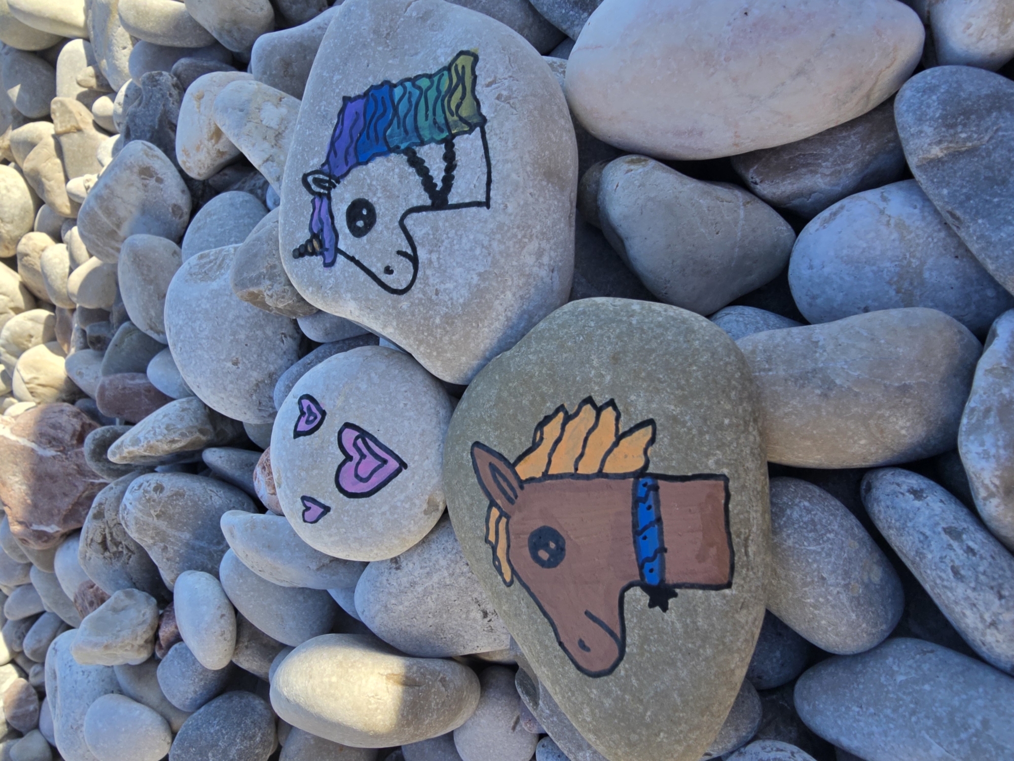 The image shows three painted rocks on a bed of smooth, light-colored pebbles. One rock features a brown horse's head with an orange mane and a blue collar. Another rock is painted with a unicorn's head, characterized by a purple horn, black eye, and a rainbow-colored mane. The third rock is decorated with three pink hearts and two simple eyes. The scene is illuminated by natural light, casting soft shadows. The location is identified as Mirište, Montenegro.