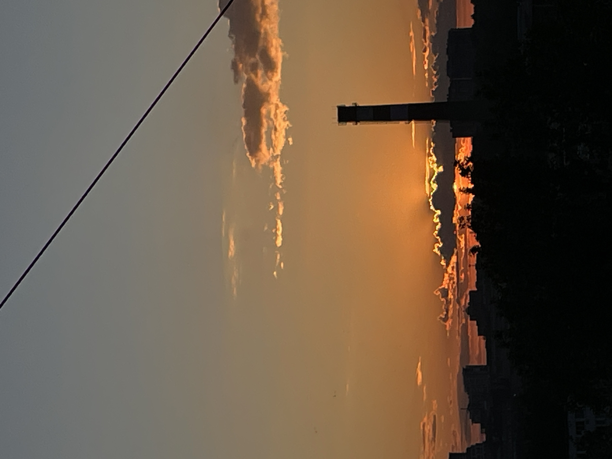 The image displays an outdoor wide shot captured at dusk or dawn, depicting an urban or semi-urban landscape. The sky occupies the upper two-thirds of the frame, exhibiting a horizontal gradient from a muted blue-grey at the very top to vibrant oranges and deep amber near the horizon. Scattered cumulus and stratocumulus clouds are present across the mid-sky, with those nearer the horizon brightly illuminated from behind by the partially visible sun, displaying distinct golden and orange edges and undersides. The sun itself is low on the horizon, partially obscured by clouds but casting a strong, golden-orange glow and some visible sunbeams through breaks in the clouds.
A dark, thin utility cable or wire diagonally traverses the upper portion of the frame from the upper-left corner towards the upper-right.
A prominent, tall, slender industrial smokestack, predominantly dark in silhouette, stands vertically slightly to the right of the central axis, extending into the mid-sky. Its upper section appears to have alternating light and dark horizontal bands, though heavily silhouetted against the bright sky.
A distant cityscape forms a dark, silhouetted skyline below the vibrant sky, composed of indistinct buildings of varying heights. Below the distant buildings, a dense, dark mass of silhouetted trees and foliage occupies the lower portion of the image, obscuring the immediate foreground and ground level. No human figures, animals, or discernible actions are visible within the frame. The scene is lit from behind by the setting or rising sun, creating strong silhouettes and warm ambient light. The location is in the city of Unknown, Unknown.