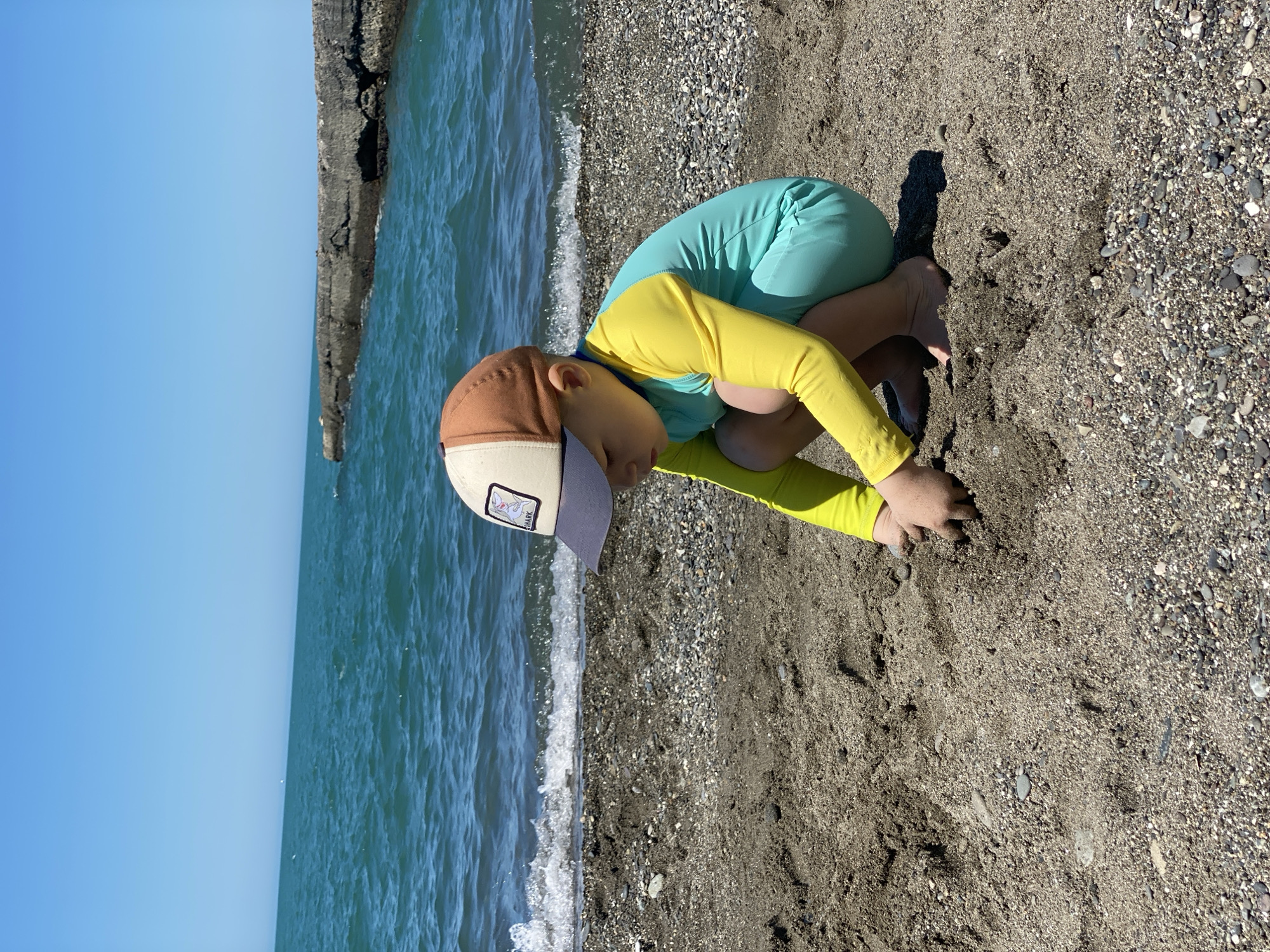 A young child, observed from a slightly elevated perspective, squats on a dark pebble and sand beach under a clear azure sky. The child wears a beige and brown baseball cap with a dark blue brim featuring a rectangular patch that partially displays the word "SHARK." Their attire also includes a long-sleeved rash guard, yellow on the sleeves and upper torso, and light blue (teal) on the lower torso and shorts. The child is barefoot and engaged in digging into the damp, dark sand with both hands. In the midground, the turquoise waters of the Black Sea exhibit gentle waves breaking on the shore, consistent with the coastal environment of Sochi, Russia. To the right, a weathered, dark concrete or rock breakwater extends into the sea.