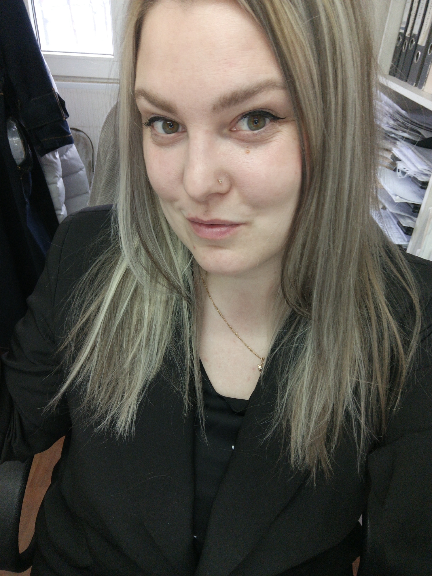 A fair-skinned woman is the primary subject of this medium-shot photograph. She has light brown eyes, and her hair is dyed with blonde and grey streaks. She is wearing a black blazer over a black top and a gold chain necklace. A small stud is visible in her left nostril. The background suggests an office environment, with a coat hanging on a chair, a window, and shelves stacked with documents. The location in the city of Noviy, Russia is not discernible from the image.