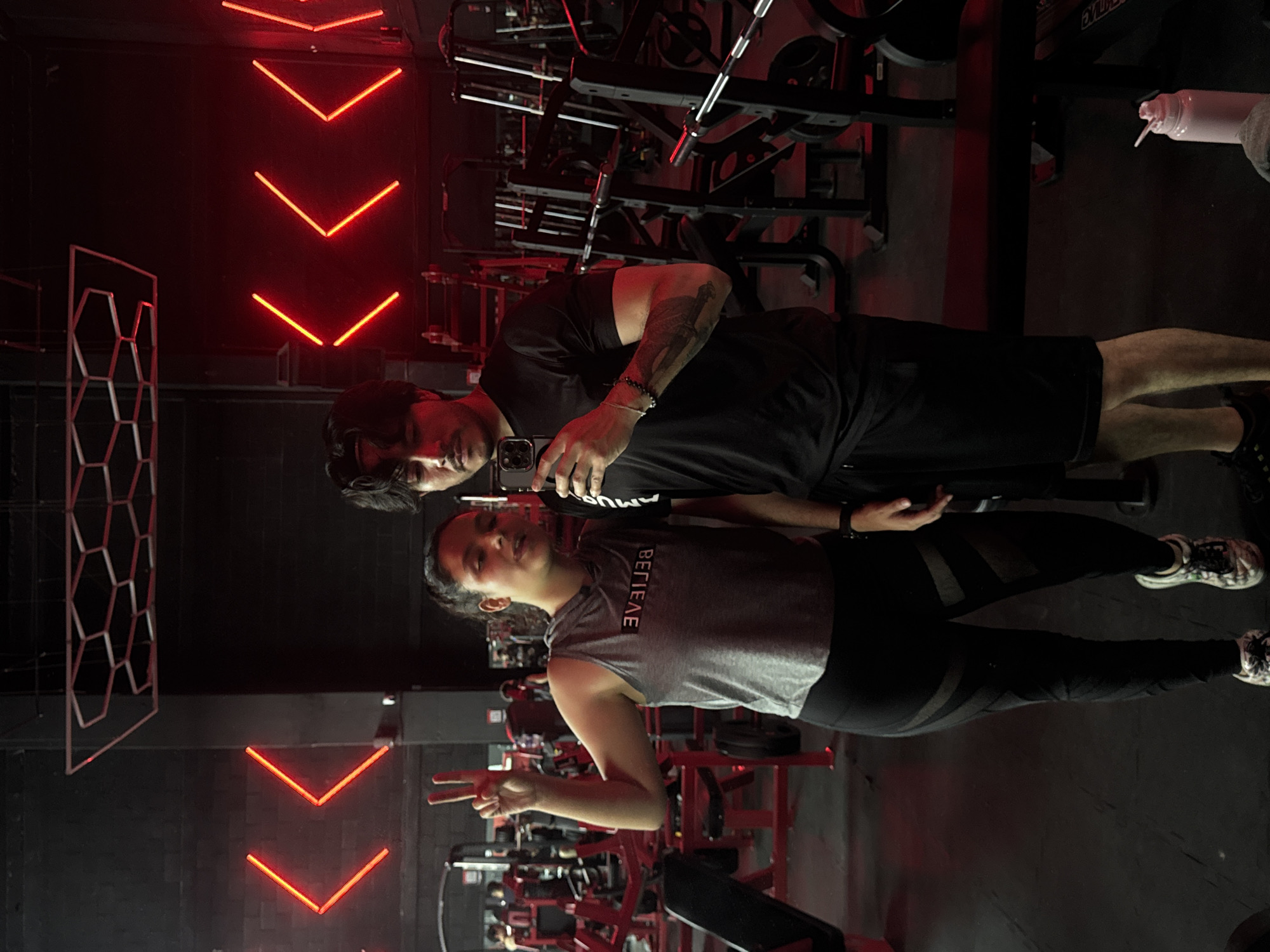 A man and a woman take a selfie in a gym. The man, wearing a black t-shirt and shorts, holds the phone. The woman wears a grey tank top with the word "BELIEVE" printed on it, and black leggings, she is gesturing with a peace sign. The gym features red neon lights and a honeycomb-shaped metal structure on the wall. Various exercise machines are visible in the background. The location is in Puebla, Mexico.