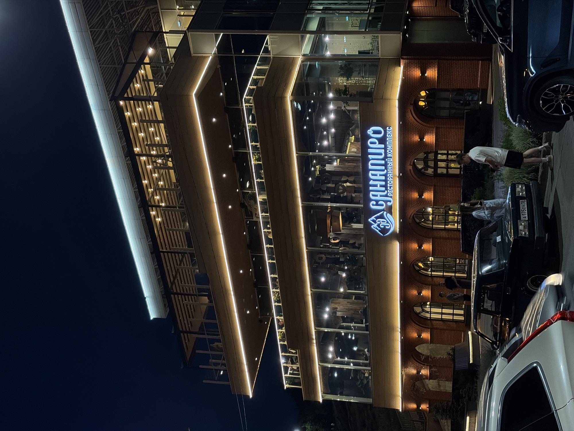 The image depicts a modern building with illuminated features at night. The building is identified by a sign in Russian that reads "САНАПИРО РЕСТОРАННЫЙ КОМПЛЕКС", indicating that it is a restaurant complex named "Sanapiro". The architecture includes a multi-tiered design with large glass windows that reflect interior lighting, suggesting a restaurant or entertainment venue with multiple levels.
Several people are visible on the sidewalk in front of the building, including a person wearing a white shirt and dark shorts. The area appears to be a parking zone or a street with multiple cars parked along the curb, including a white SUV and a black sedan.
Based on the sign and the cityscape, the location is likely in Izhevsk, Russia. The image captures a modern, urban scene at night, with a focus on the illuminated architecture of the Sanapiro restaurant complex.